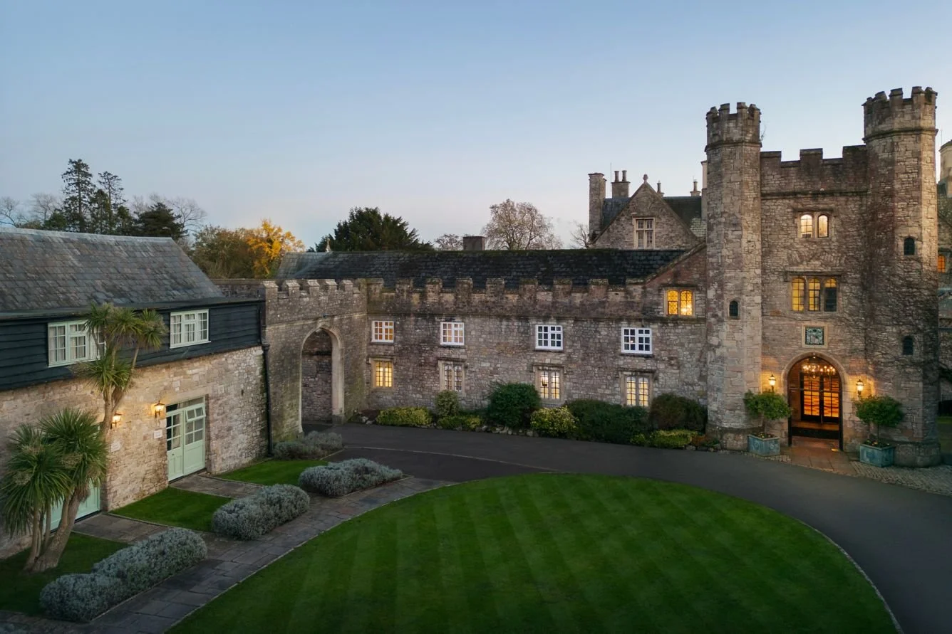 A stone castle-like building with a courtyard and manicured lawn, illuminated windows, and an arched entrance, during dusk.