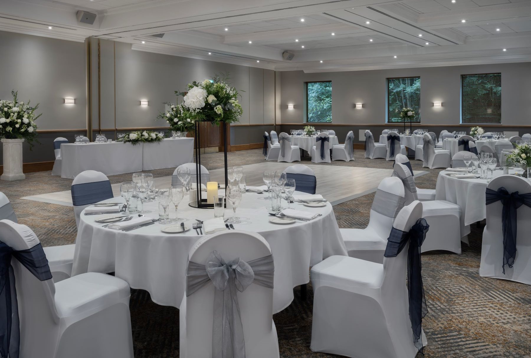 Elegant banquet room decorated for a wedding reception with round tables covered in white tablecloths, set with glassware, silverware, and white napkins. Chairs are draped in white fabric with gray sashes. Centerpieces feature white flowers and candles. The room has gray walls, wooden trim, and large windows with blinds, illuminated by ceiling lights.