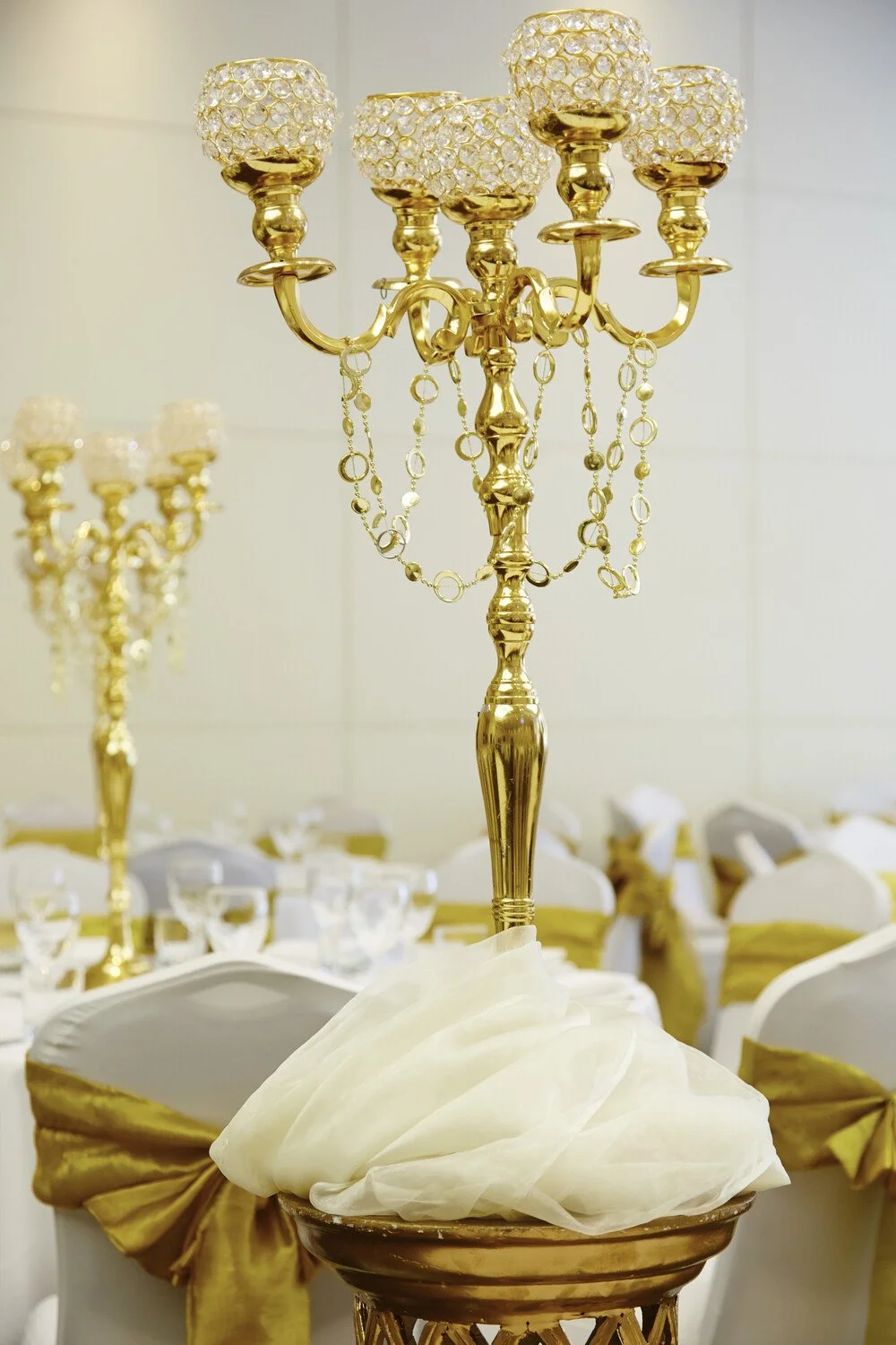 Gold chandelier with crystal accents and dangling chains, decorated with white fabric, in a decorated banquet hall with white and gold chairs and tables set for an event.