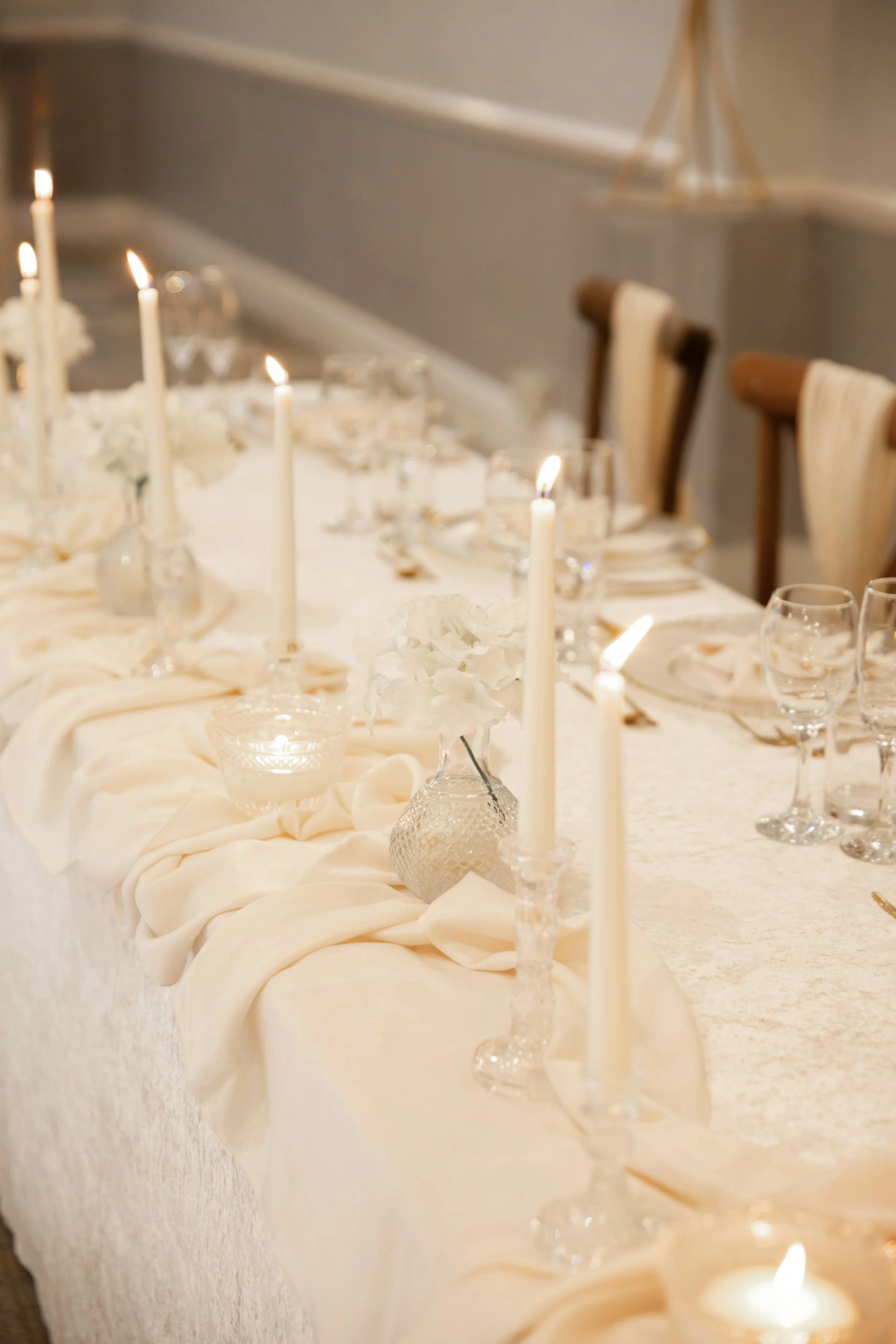 A long dining table decorated with white candles, flowers, glassware, and cream-colored tablecloth for a formal event.