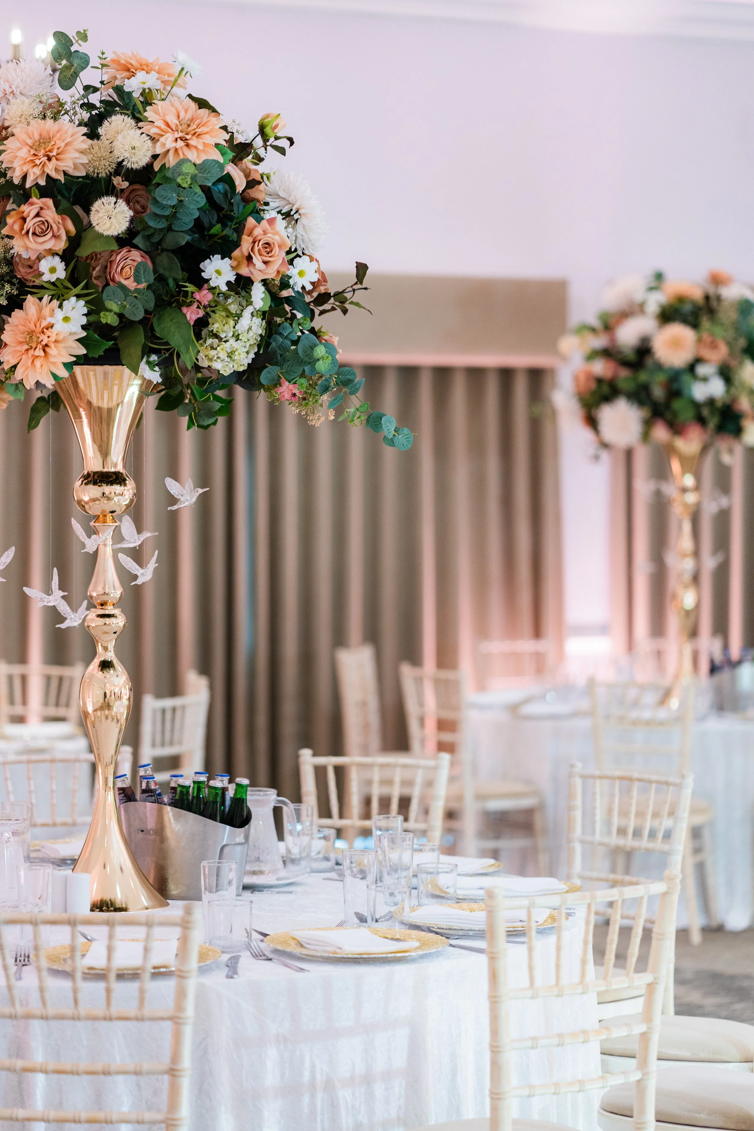 Elegant banquet table with floral centerpiece in a gold vase, set with plates, glasses, and utensils, surrounded by chairs, with another floral arrangement in the background, in a decorated event hall.