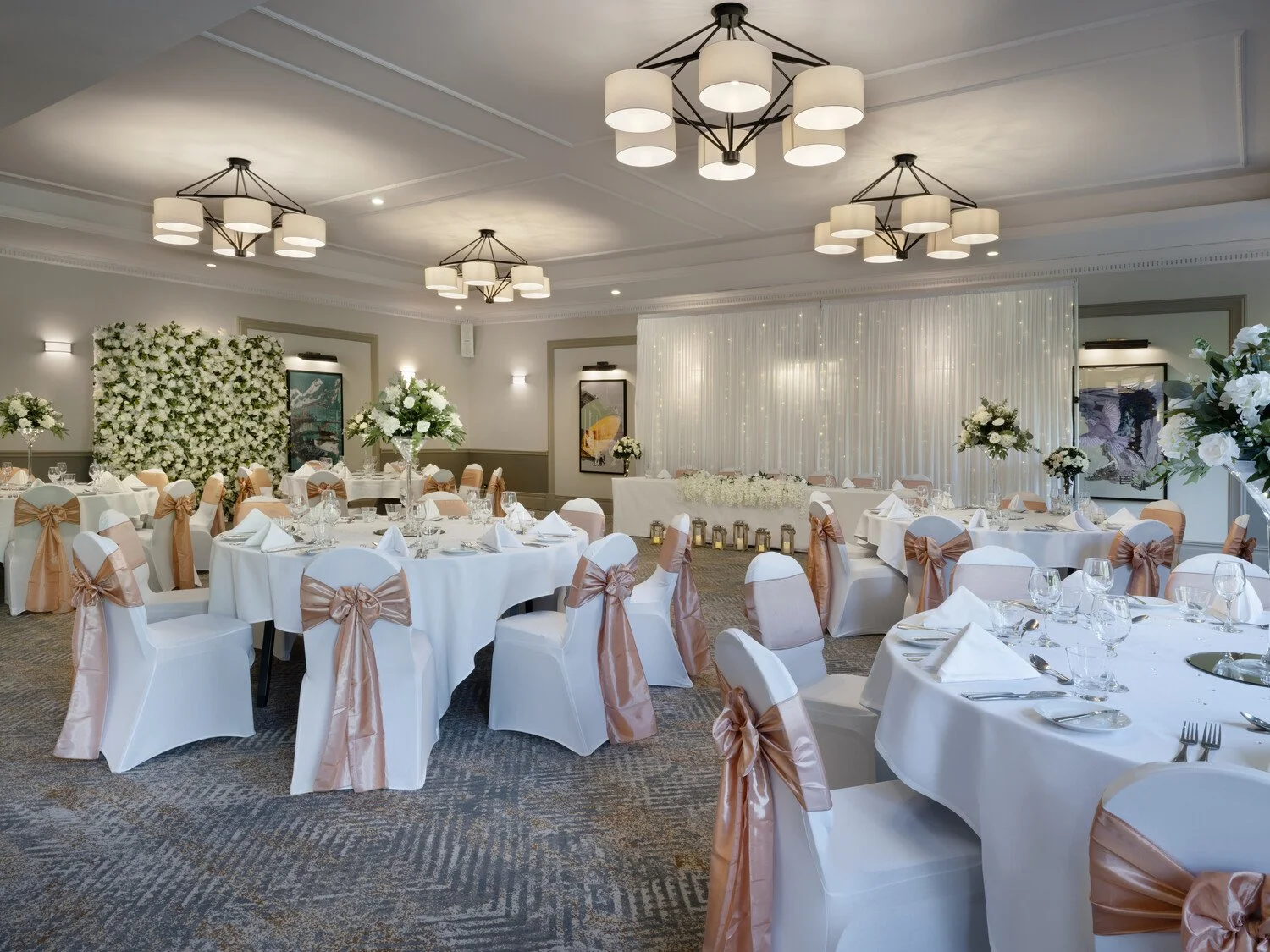 Elegant banquet hall decorated for a wedding reception with round tables, white tablecloths, floral centerpieces, and chairs with peach-colored sashes. There are floral arrangements on the walls and a backdrop for photos or a ceremony.