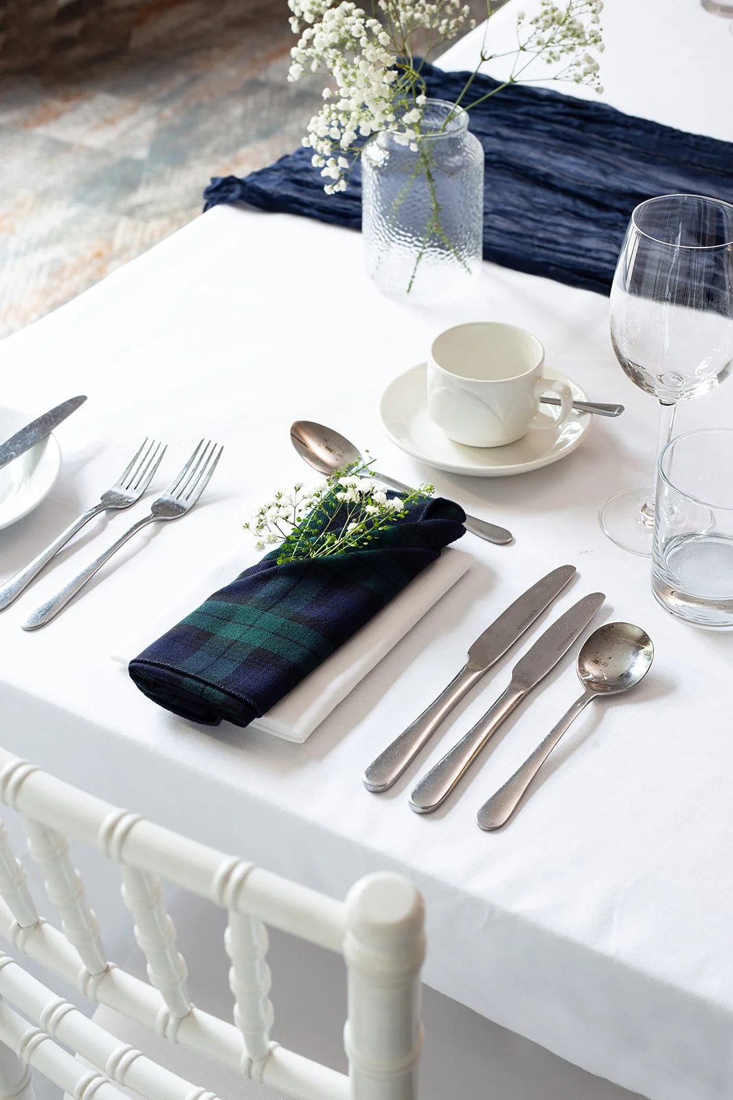 A formal dining table set with silverware, a white coffee cup on a saucer, two wine glasses, a clear glass water glass, a white cloth napkin with a dark plaid napkin, small white flowers, and a glass vase with white flowering branches. The table has a white tablecloth and a dark blue runner.