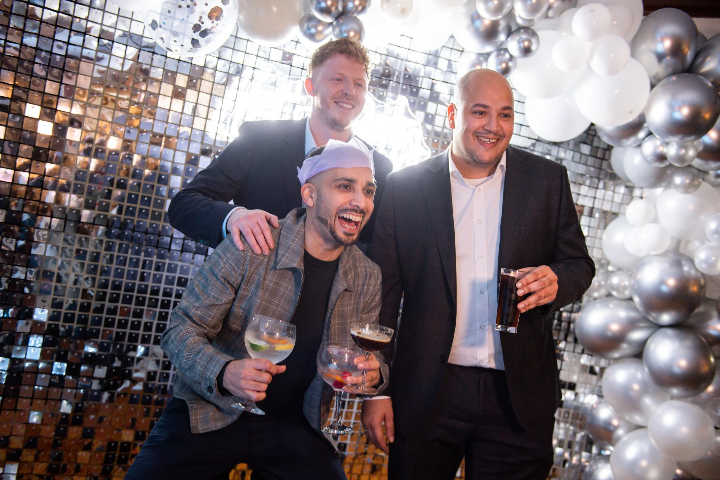 Three men at a celebration, smiling and holding drinks, in front of a decorated background with balloons and a mirror tile wall.