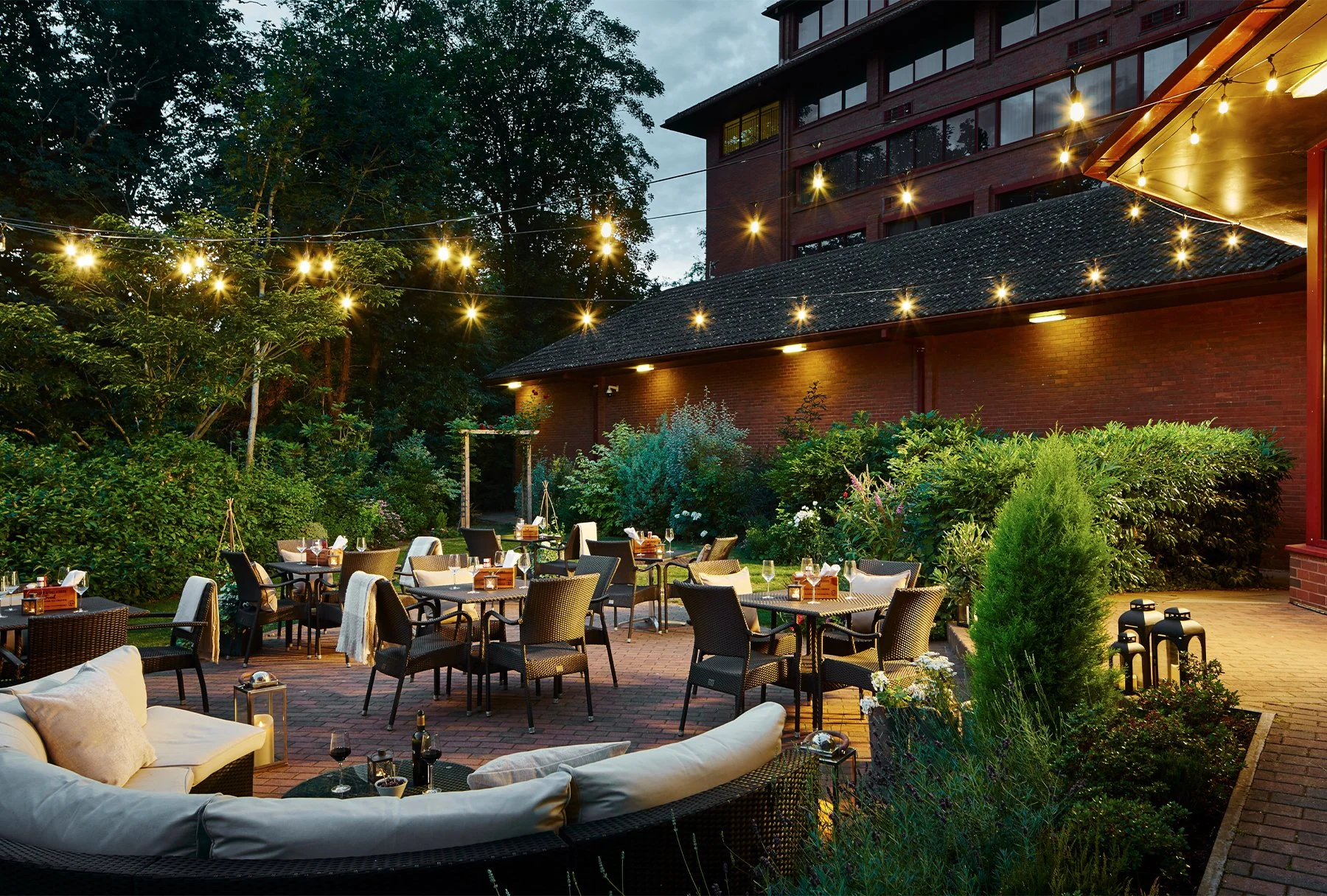 Outdoor restaurant patio with tables set for dining, string lights hanging overhead, surrounded by lush greenery and a brick building in the background during evening.