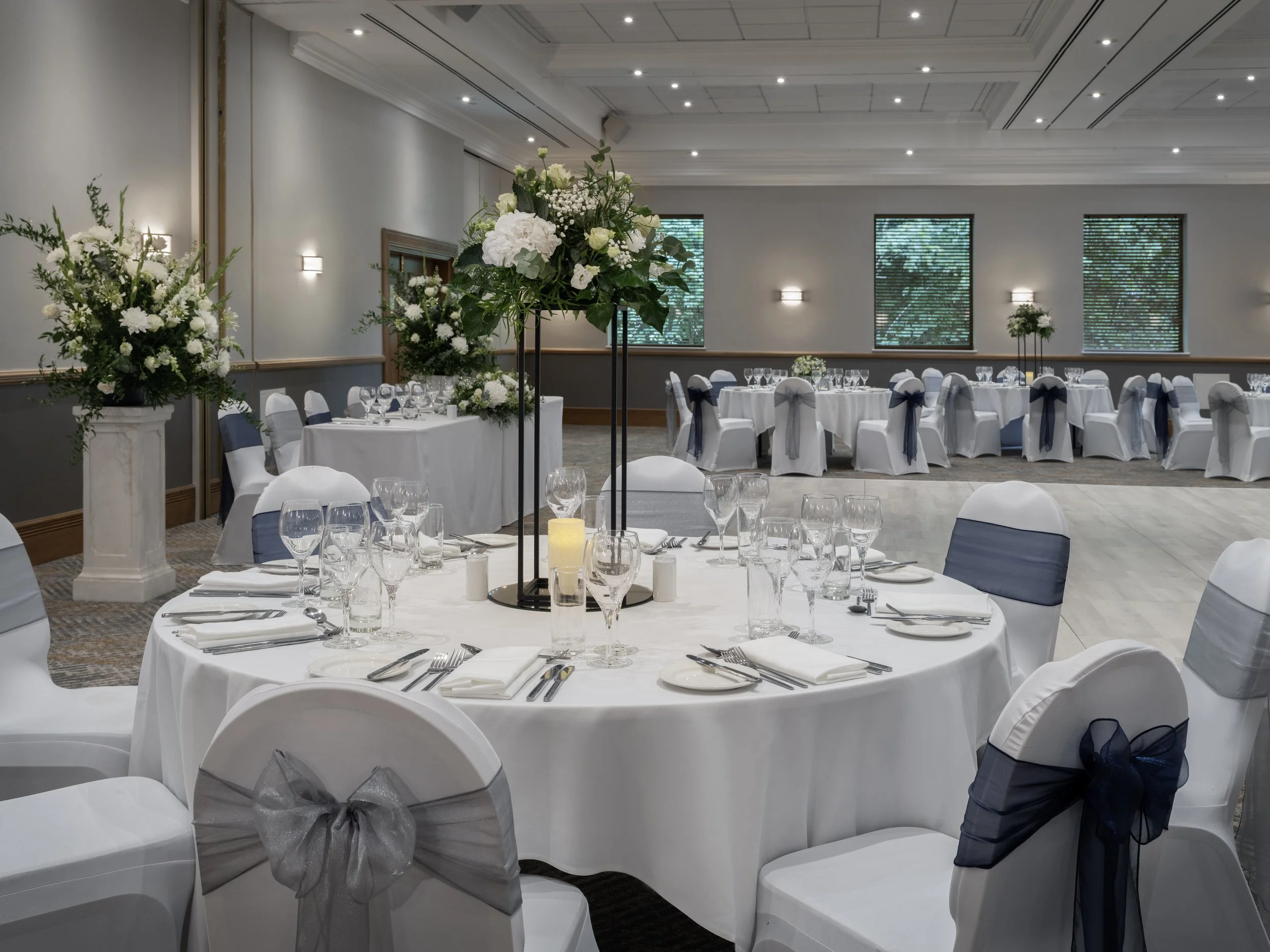 Elegant event space decorated for a banquet with round tables covered in white tablecloths, adorned with floral centerpieces, and chairs decorated with navy and gray bows.