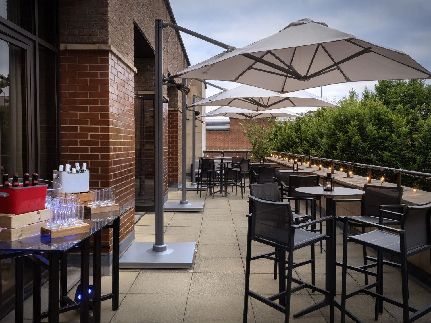 Outdoor rooftop patio with black chairs and tables, white umbrellas, string lights, and trees in the background.