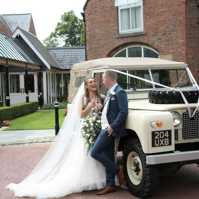 Bride and groom celebrating next to a vintage vehicle decorated with white ribbon, in front of a red brick building with a white-framed window.