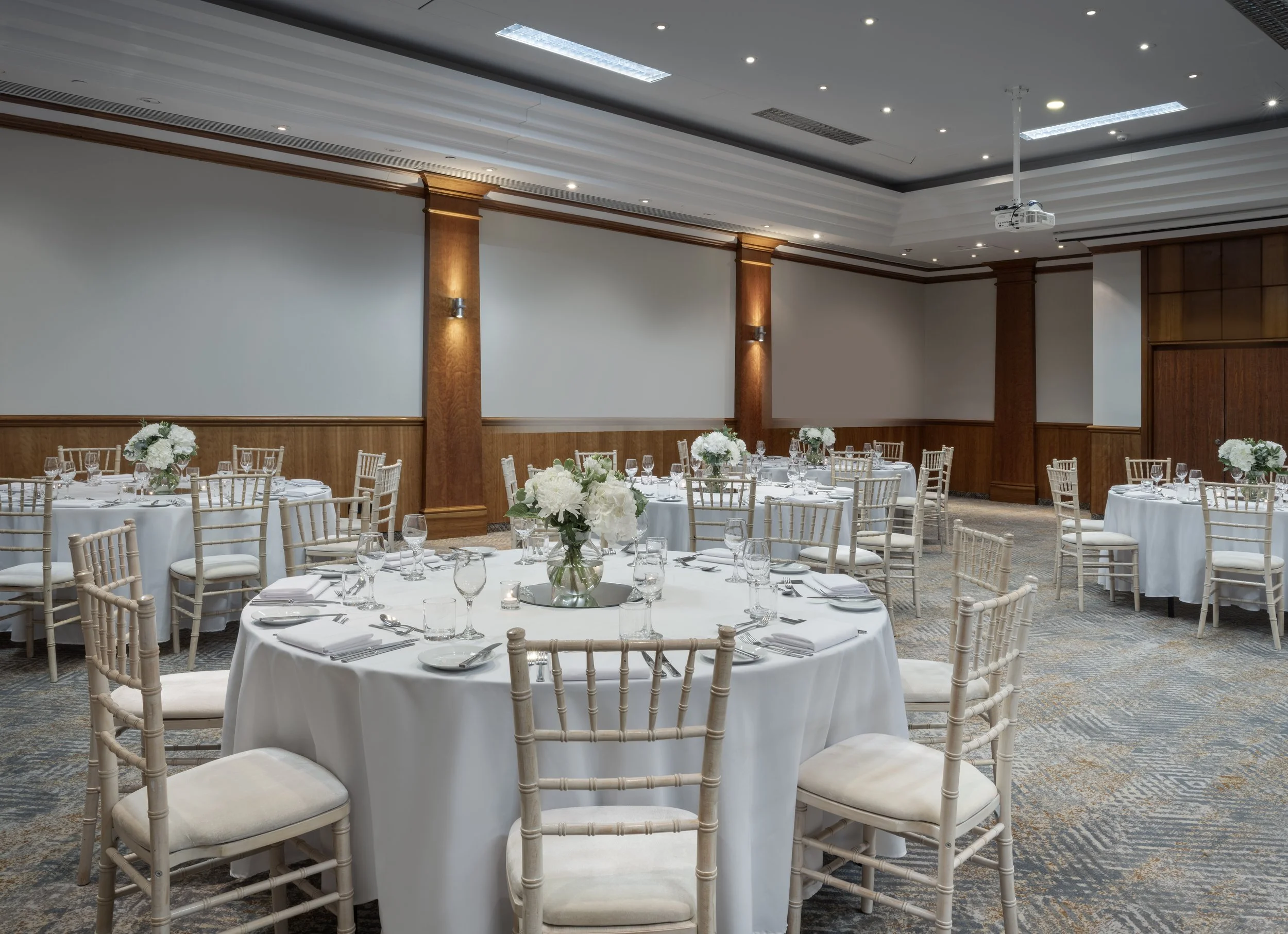 Banquet hall with round tables covered with white tablecloths, set with glassware, silverware, napkins, and floral centerpieces, surrounded by light-colored chairs. The room has a modern ceiling with recessed lighting, wood panel accents, and a mounted projector.
