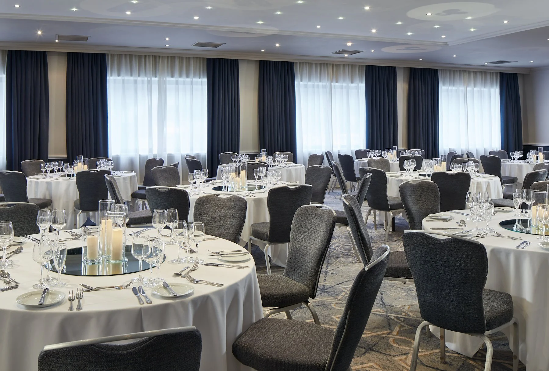 Elegant banquet room with round tables covered in white tablecloths, set with glassware, silverware, and centerpieces featuring candles. The room has large windows with sheer curtains and dark drapes, and a patterned carpet.