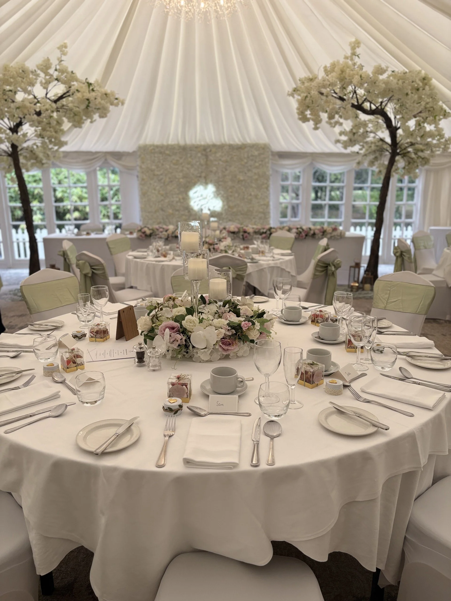 Round banquet table set for a wedding reception with white tablecloth, floral centerpiece, candles, and place settings, surrounded by white chairs with decorative bows. In the background, there are large windows, a textured wall, floral arrangements, and trees within a decorated tent.
