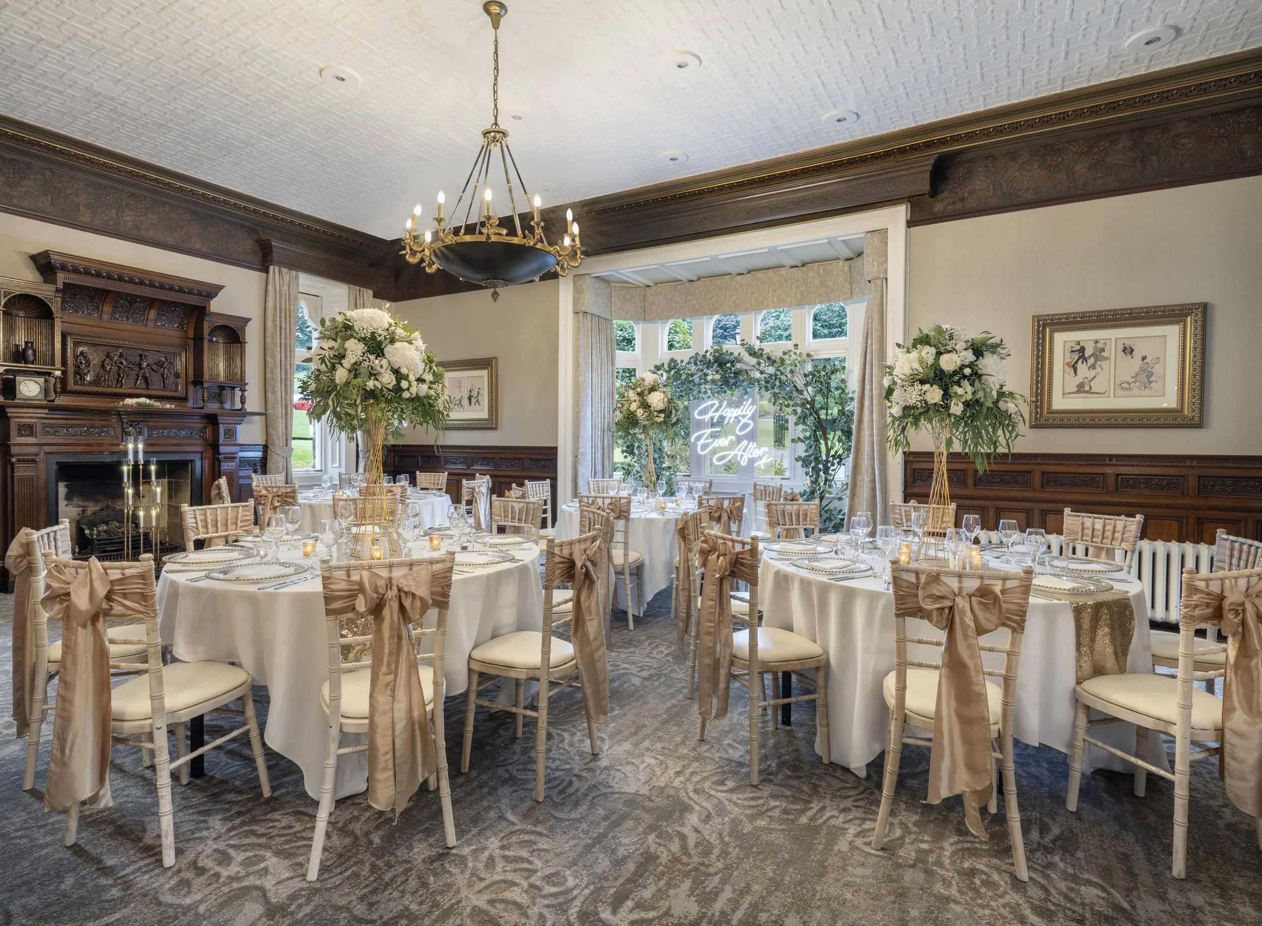 Elegant banquet hall with round tables covered in white tablecloths, decorated with floral centerpieces, and tied with beige sashes on chairs. The room features a large window with a neon sign reading "Happily Ever After," ornate woodwork, framed artwork, and a chandelier hanging from a decorated ceiling.