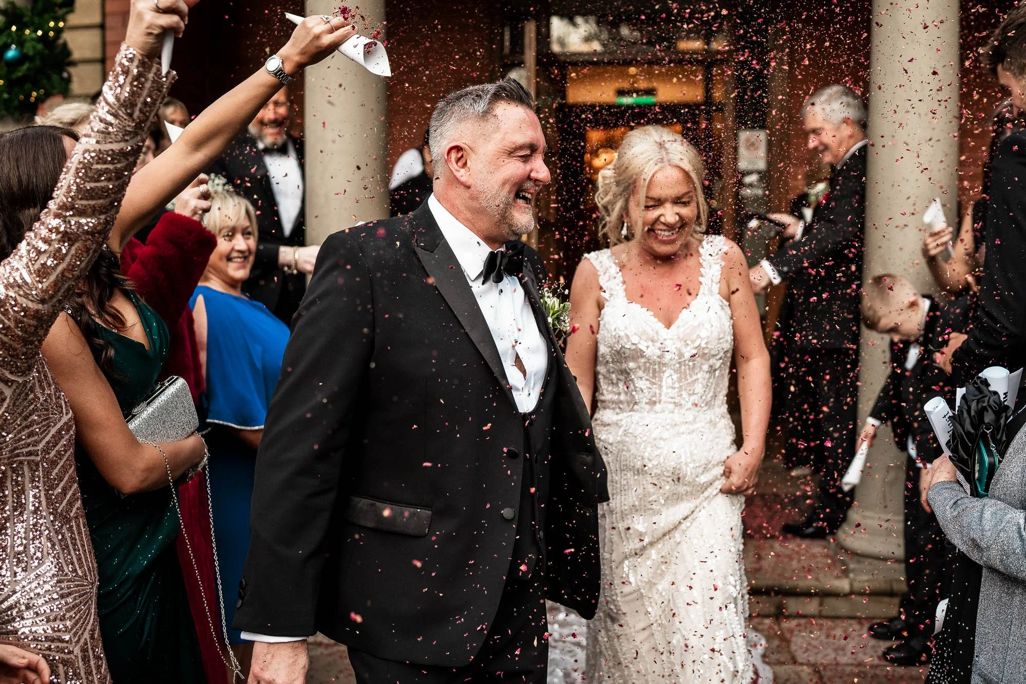 A group of people celebrating a wedding outside, with some throwing confetti. The bride and groom are at the center, smiling and dressed in wedding attire. The groom is in a black tuxedo with a bow tie, and the bride is in a white wedding dress. Other guests are around them, dressed in colorful clothes, cheering and participating in the celebration.