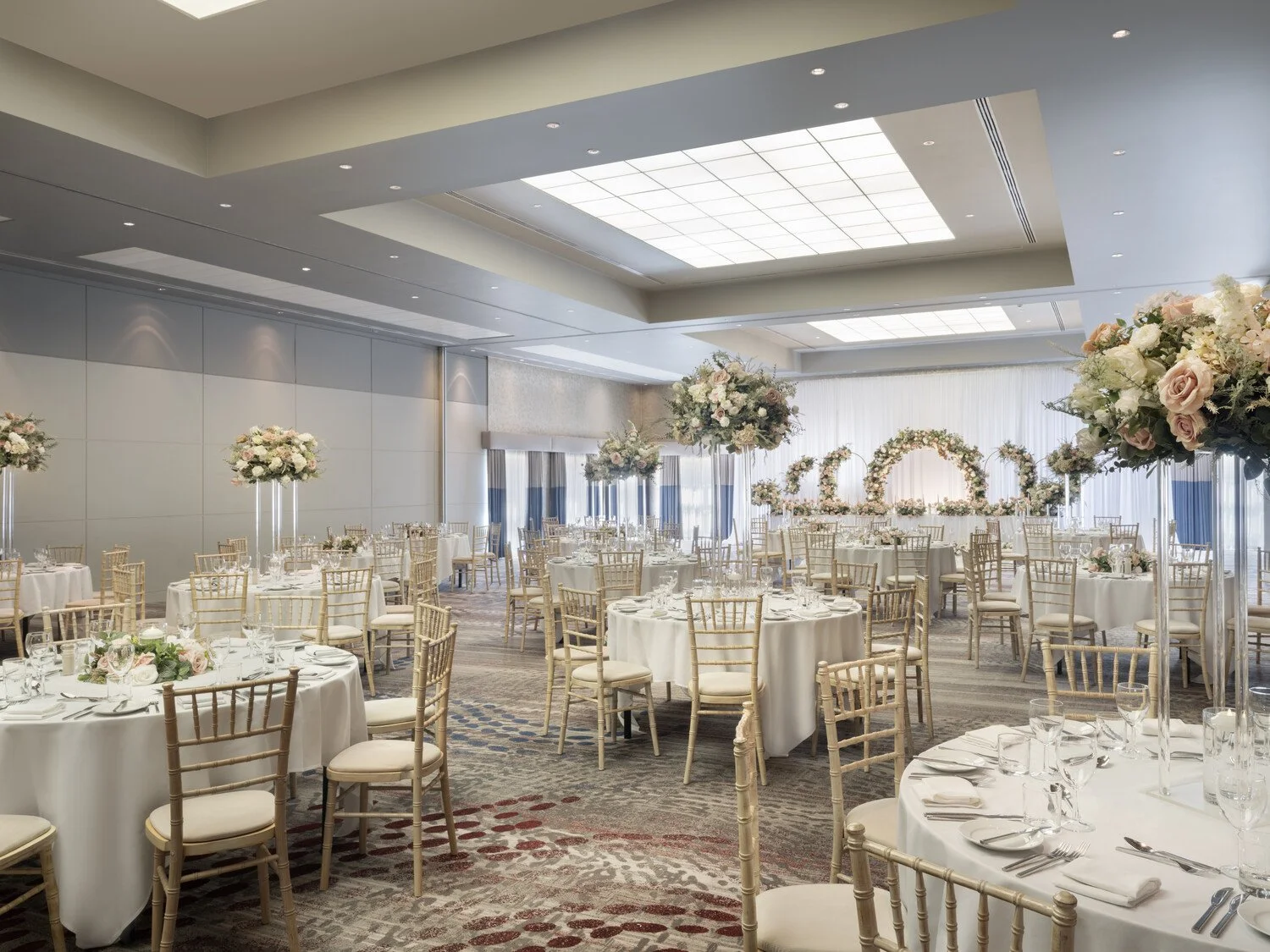 Beautifully decorated banquet hall with round tables covered in white tablecloths, elegant gold chairs, and tall floral centerpieces featuring pink and white roses and greenery. In the background, a floral arch and more floral arrangements are visible, set up for a wedding or special event.