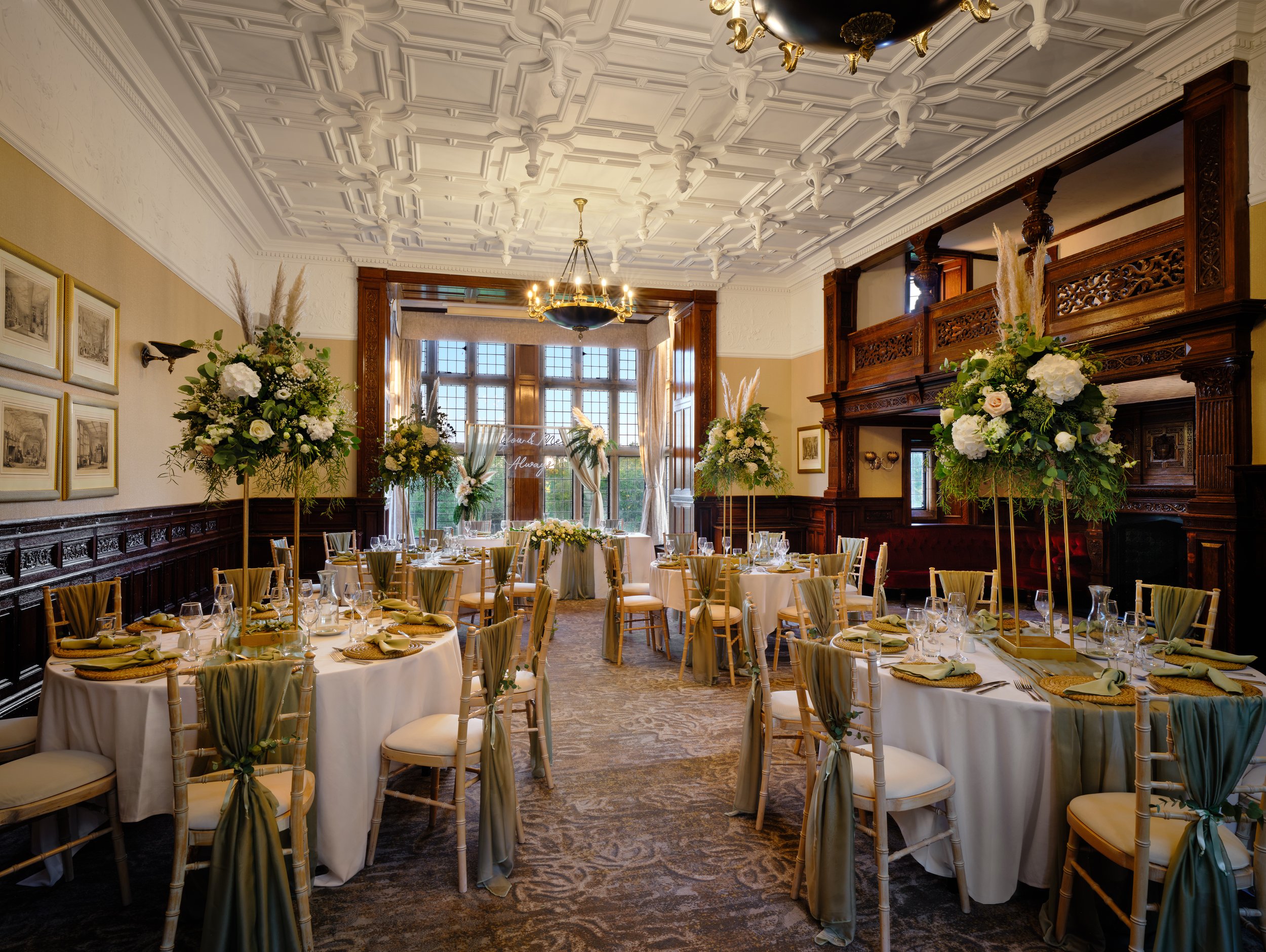 Elegant banquet hall decorated with tall flower arrangements, round tables with white tablecloths, napkins, glassware, and chairs, featuring ornate woodwork, large windows, and high ceilings with intricate molding.