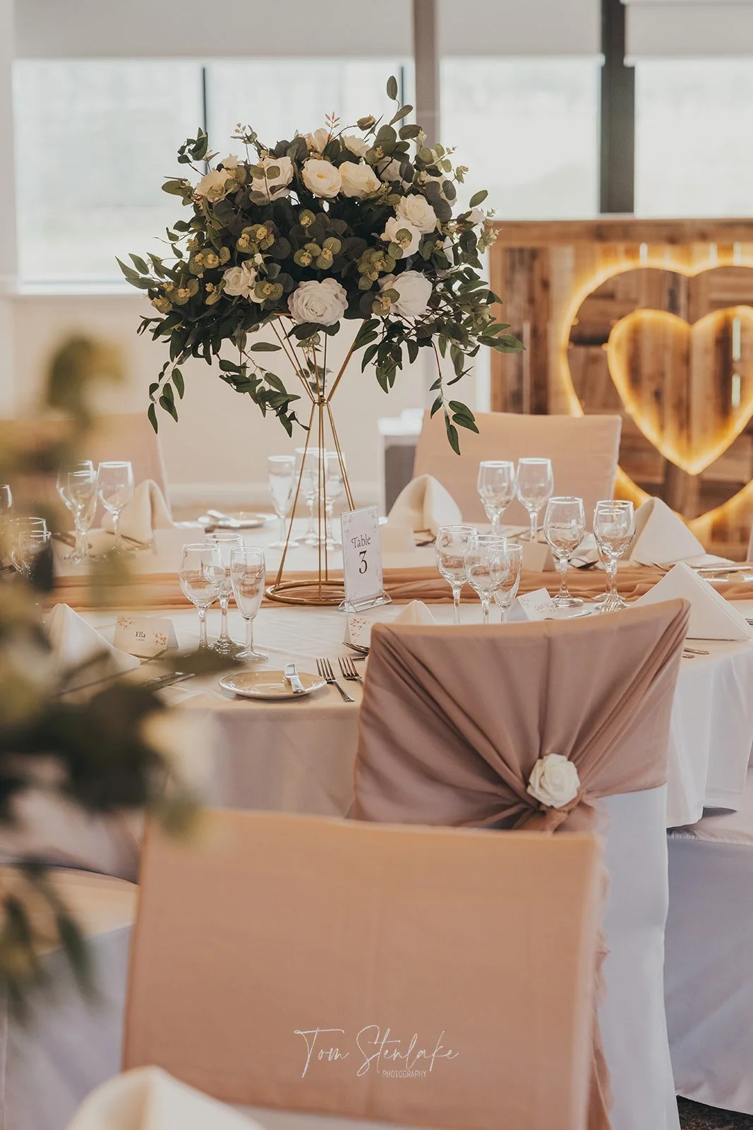 A decorated wedding reception table featuring a tall floral centerpiece with white flowers and greenery, surrounded by wine glasses, cutlery, folded napkins, and a pink chair with a tied cloth and flower detail in a cozy venue with wooden accents and heart-shaped lighting.
