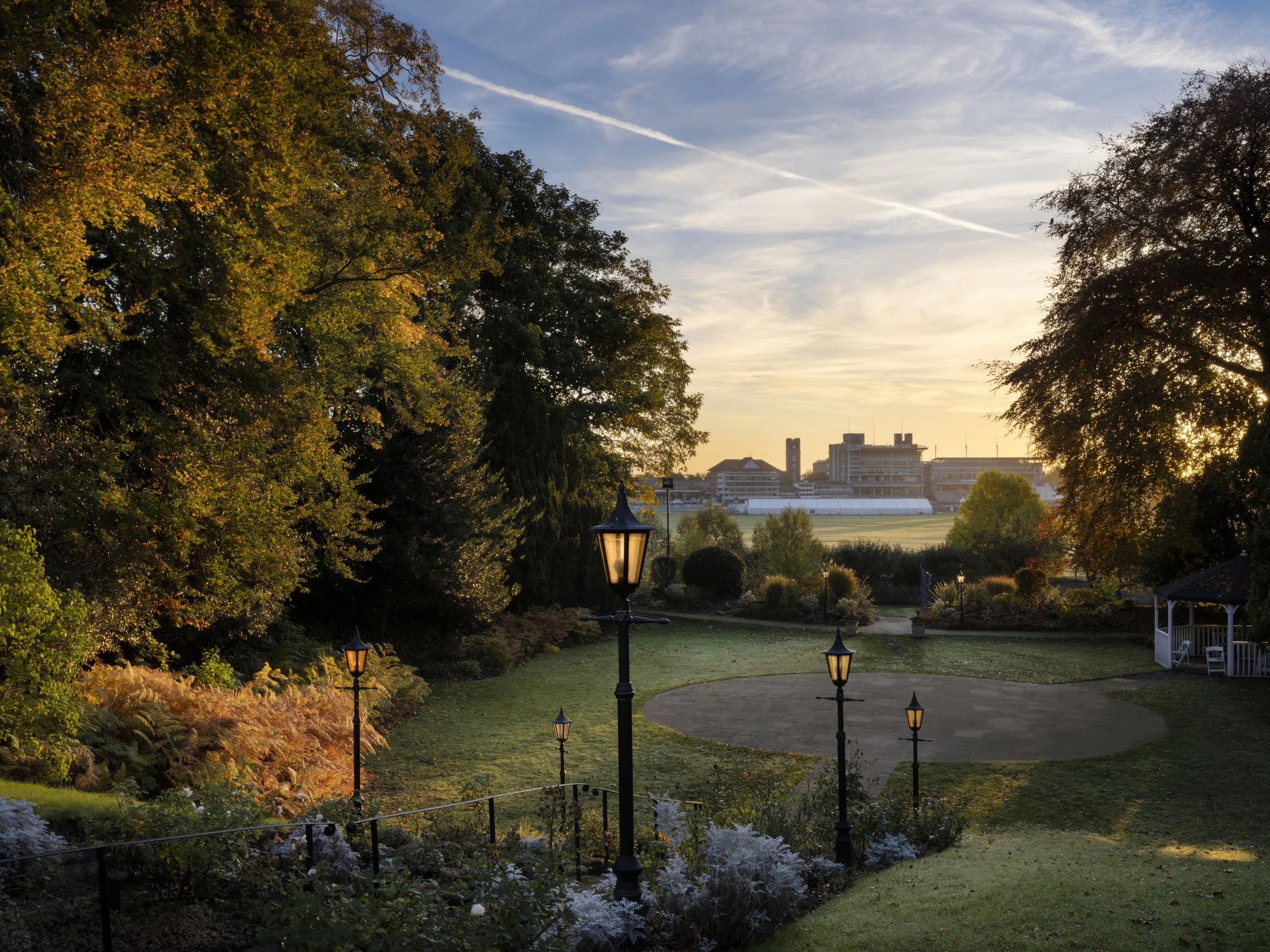 A park with trees displaying fall foliage, multiple vintage-style light posts, a walking path, and in the background, modern buildings and a field at sunset.