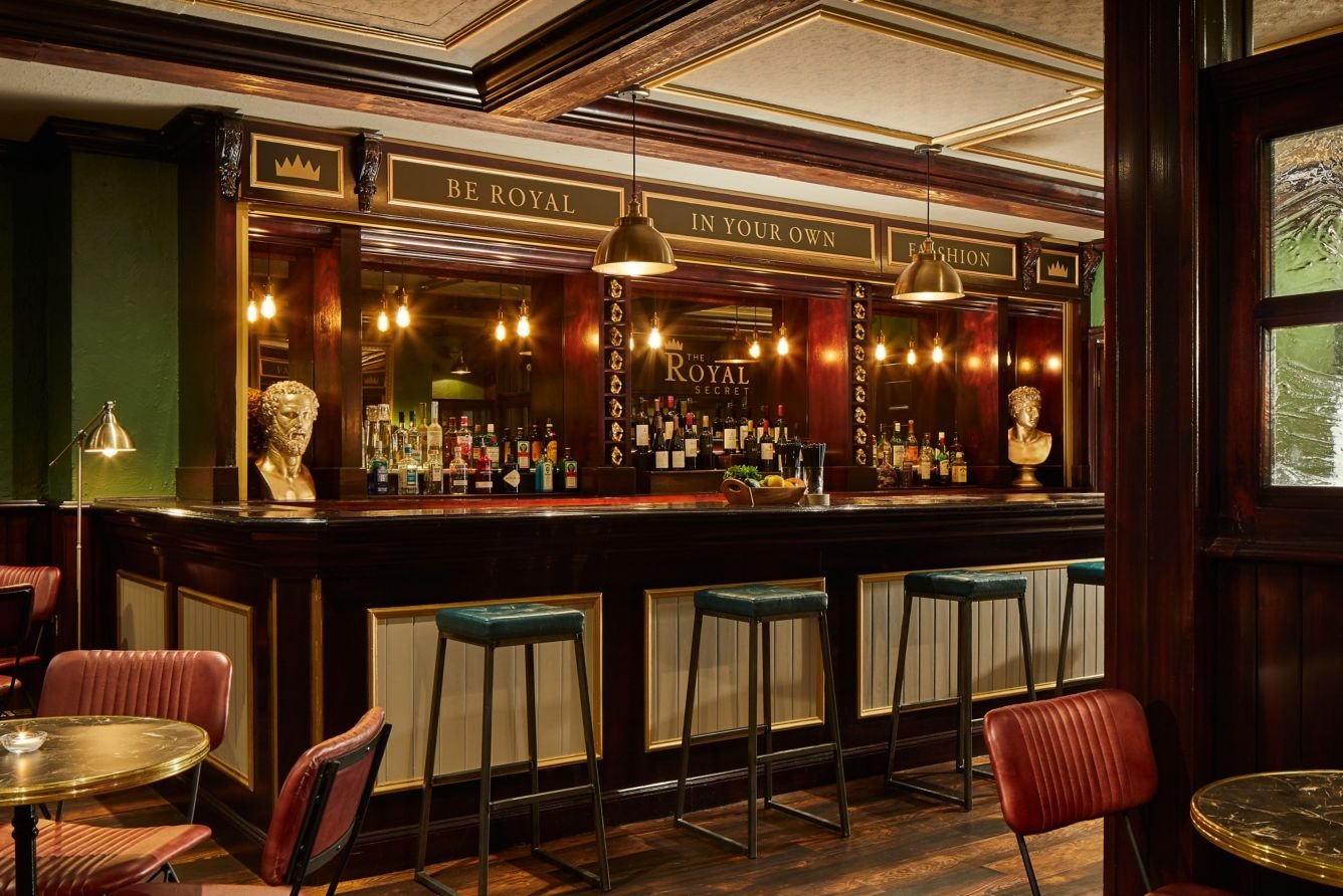 A vintage-style bar with dark wood paneling, gold accents, and a variety of liquor bottles displayed behind the counter. There are four barstools in front, and golden bust sculptures on either side of the counter. The place has warm, ambient lighting and a sign that says 'Be Royal In Your Own Fashion.'