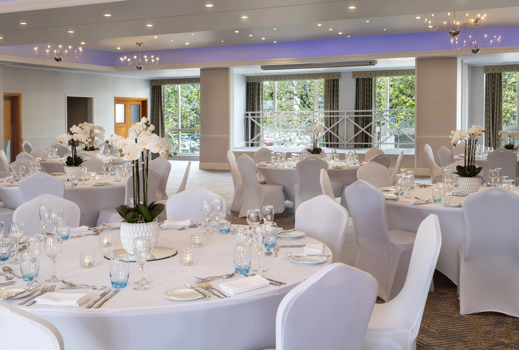 Elegant banquet hall set up for a formal event with round tables covered in white tablecloths, decorated with white orchids in vases, and set with glassware, silverware, and white napkins. Large windows reveal greenery outside, and the ceiling features chandeliers and soft purple lighting.