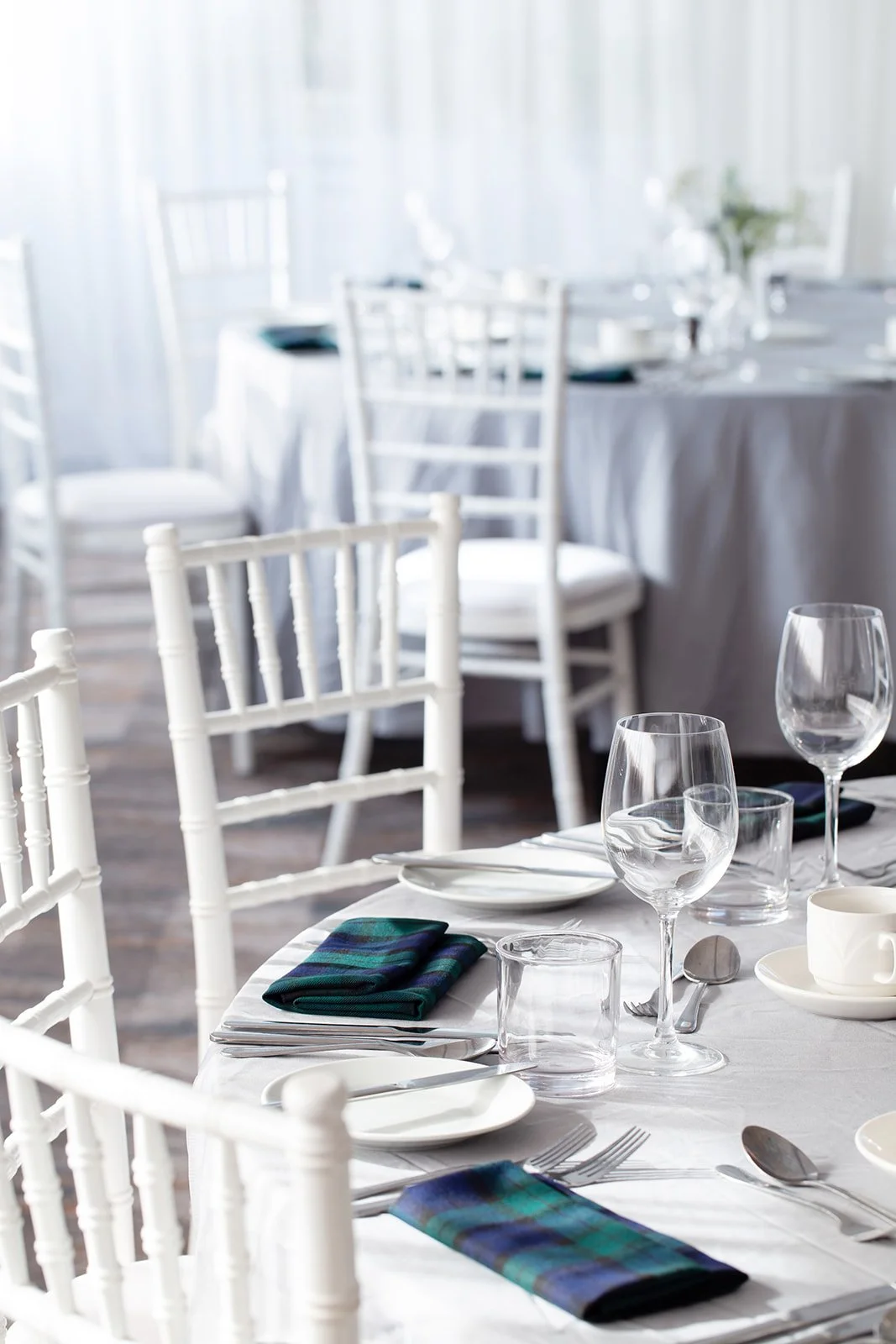 Elegant dining table set with white tablecloth, crystal glasses, white plates, napkins, and silverware in a bright, airy restaurant with white chairs and a softly lit background.