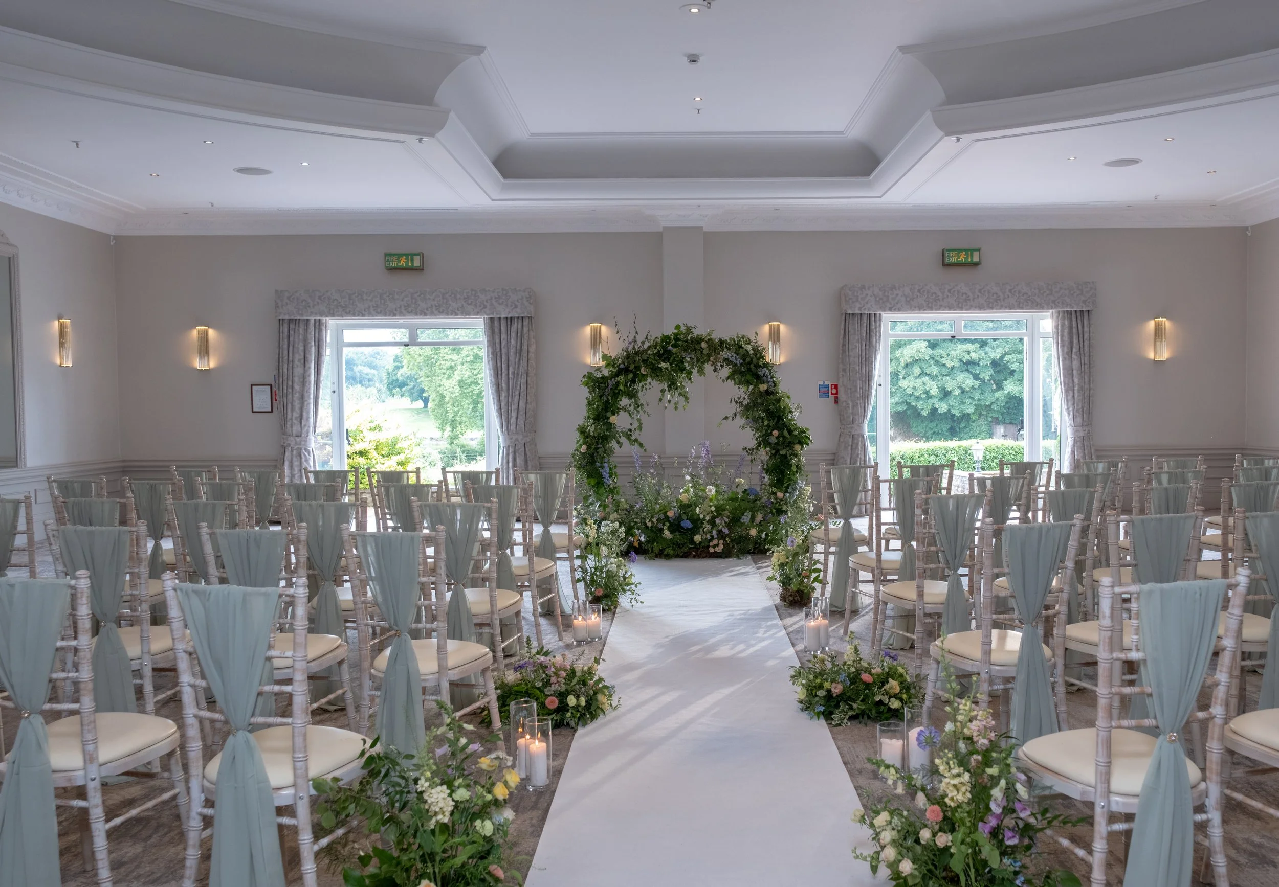 A decorated wedding ceremony space with rows of chairs covered in light fabric, an aisle with a white runner, floral arrangements, and candles, with large windows revealing greenery outside.