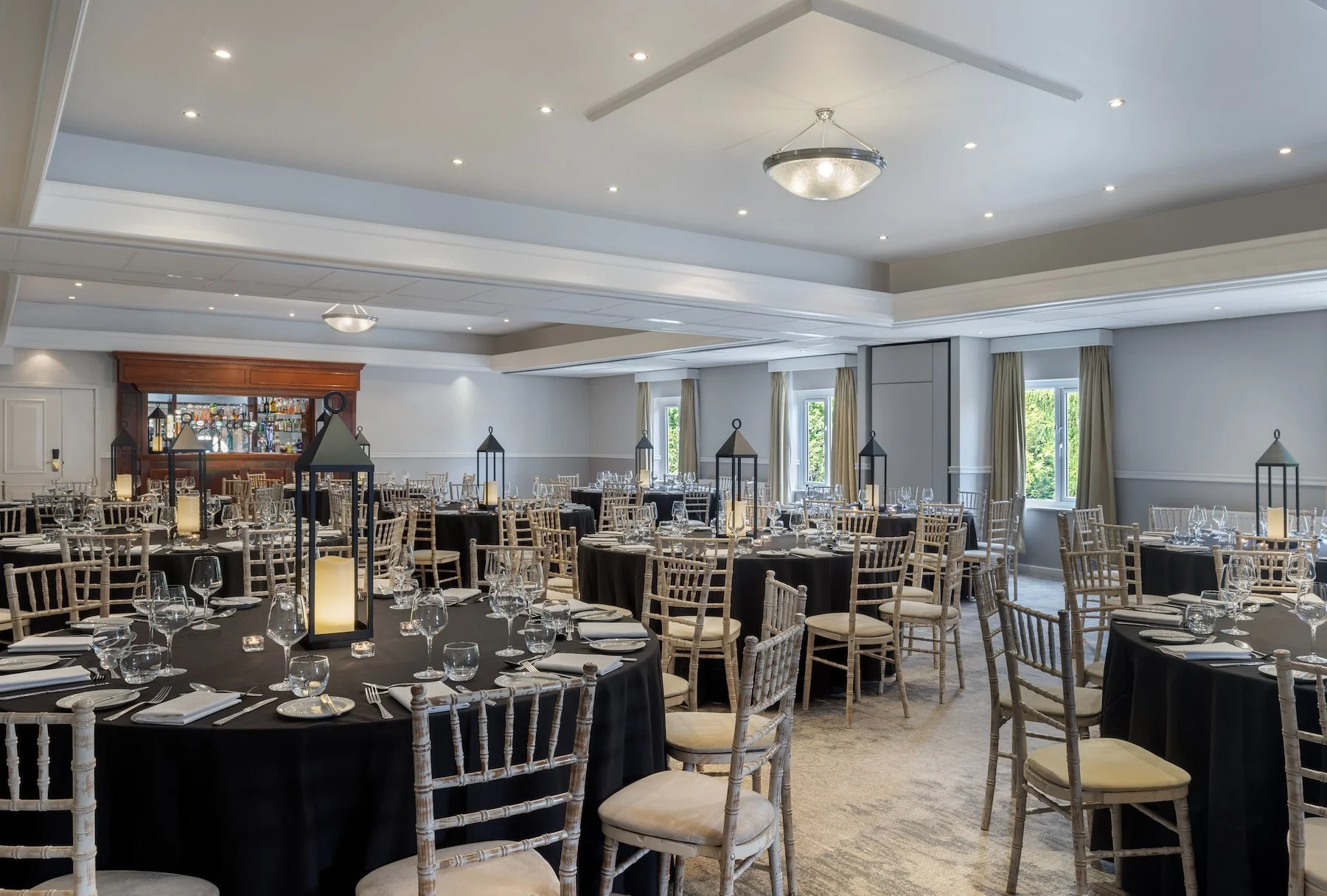 Elegant banquet hall set up for an event with round tables covered in black tablecloths, each with multiple wine glasses, silverware, and white napkins. Tall black lanterns with candles and LED candles are placed as centerpieces on each table. Light-colored wooden chairs surround the tables. The hall has large windows with beige curtains, a bar with liquor bottles in the background, and a plain ceiling with recessed lighting and modern circular light fixtures.