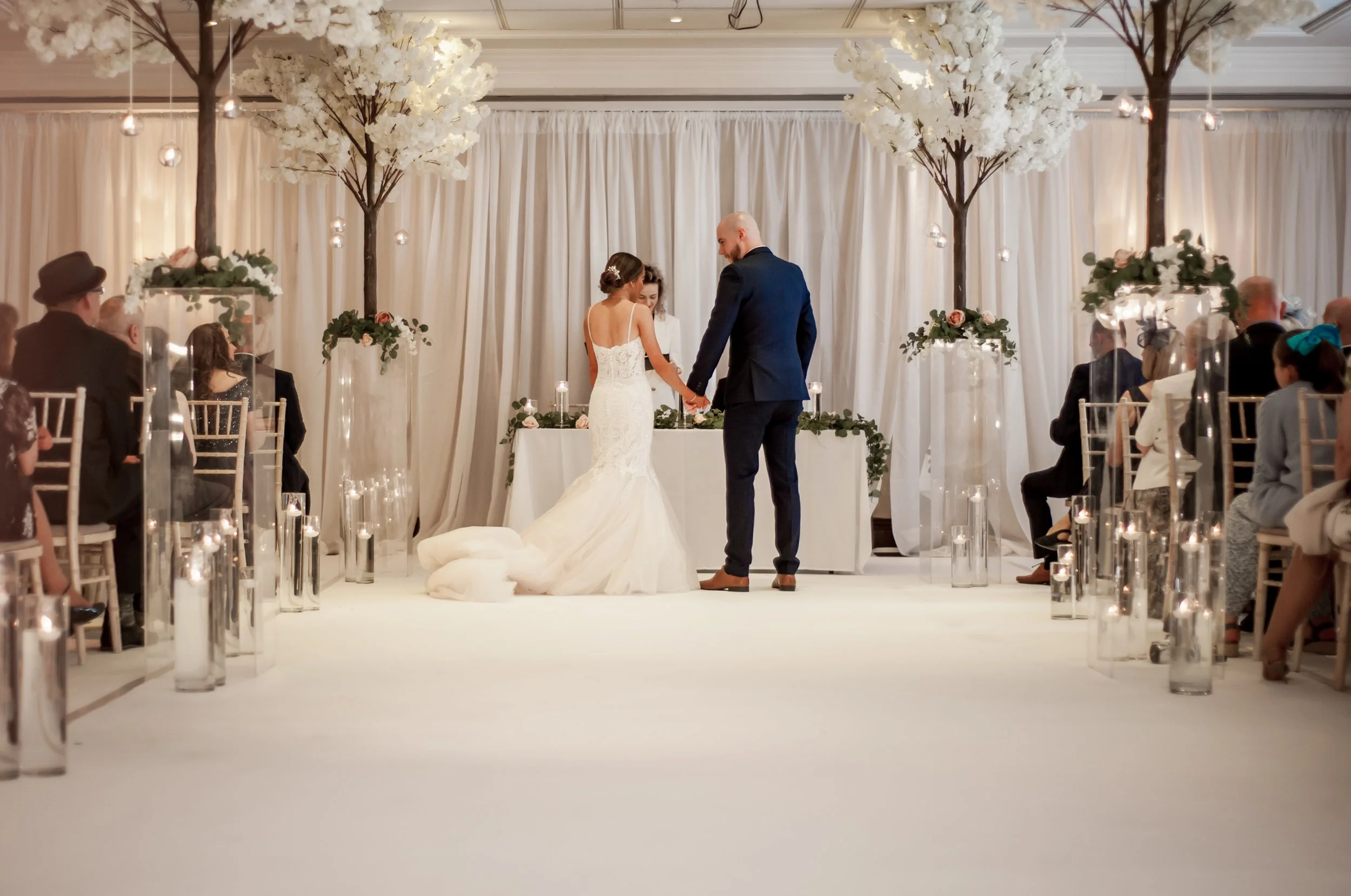 A wedding ceremony with a bride and groom holding hands, standing at an altar decorated with greenery and flowers, inside a decorated indoor venue with trees, hanging lights, and seated guests.