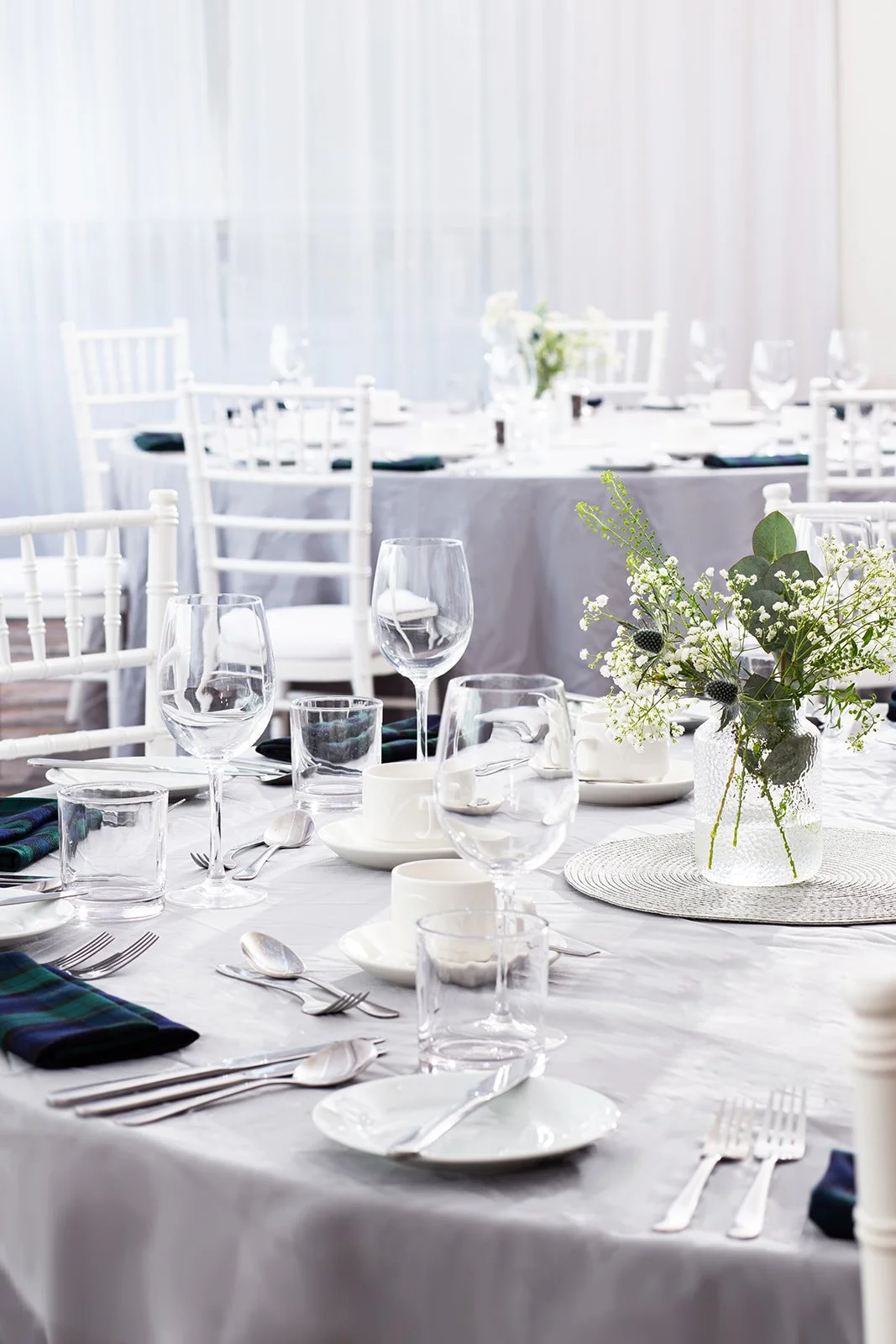 Set dining table with white tablecloth, glassware, plates, silverware, a flower arrangement, and white chairs in the background.