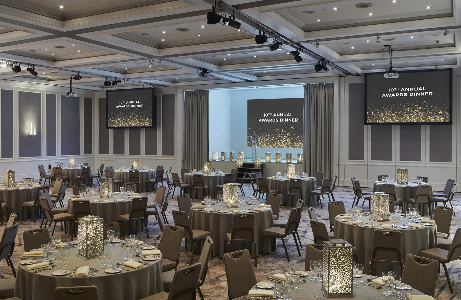 A banquet hall set up for the 10th Annual Awards Dinner, with round tables covered in champagne-colored tablecloths, each with matching napkins, glassware, and candles in decorative lanterns. The room has taupe walls with white trim, and a stage at the front with large screens displaying the event name and a starry graphic.