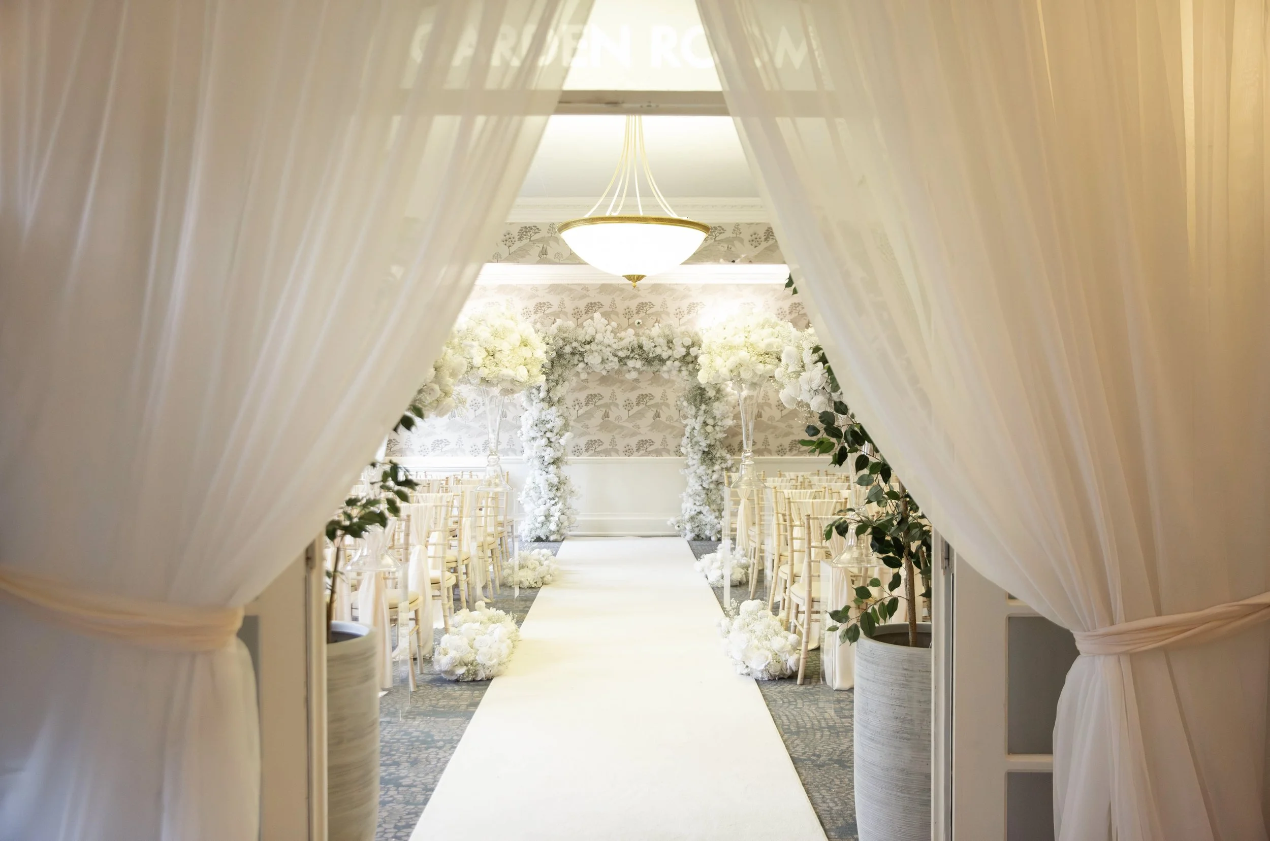 A wedding ceremony setup seen through an opening with white curtains. The aisle is decorated with white flowers and leads to a floral arch, with chairs on either side for guests.