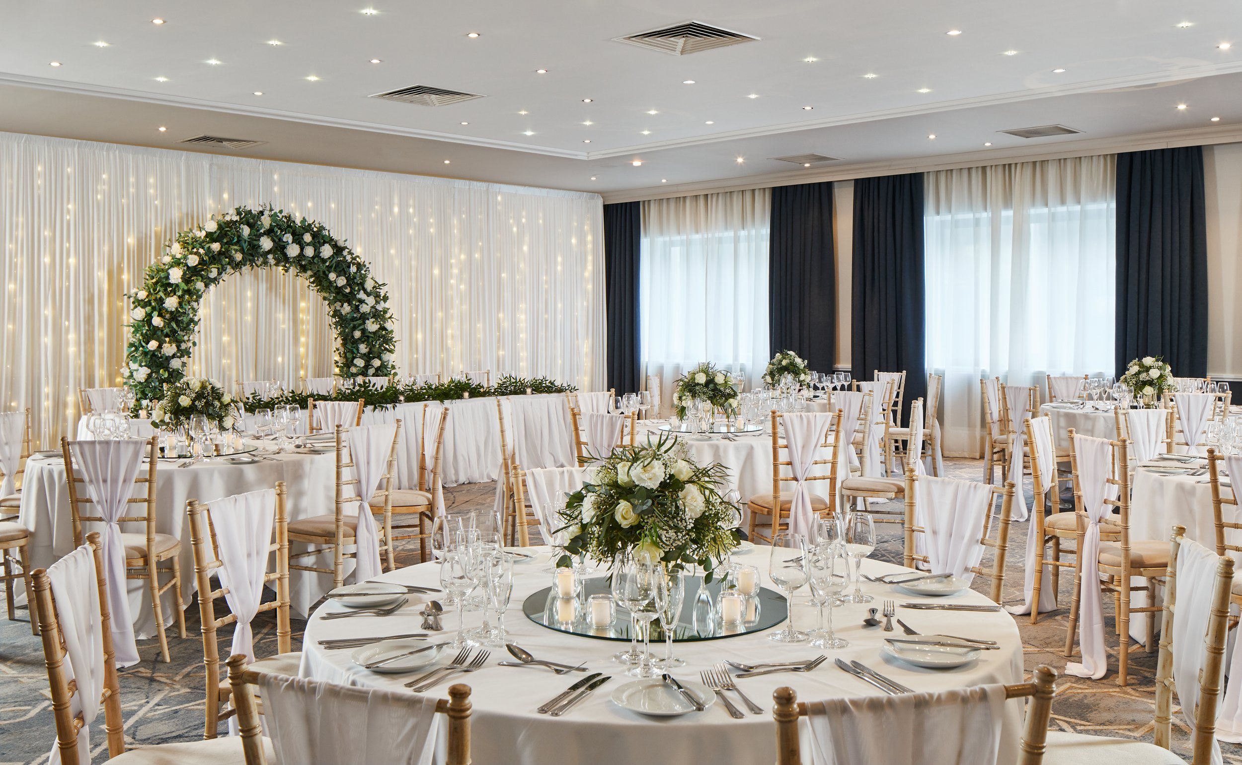 Wedding reception room decorated with white and navy curtains, a floral arch, and elegant table settings with floral centerpieces and candles.
