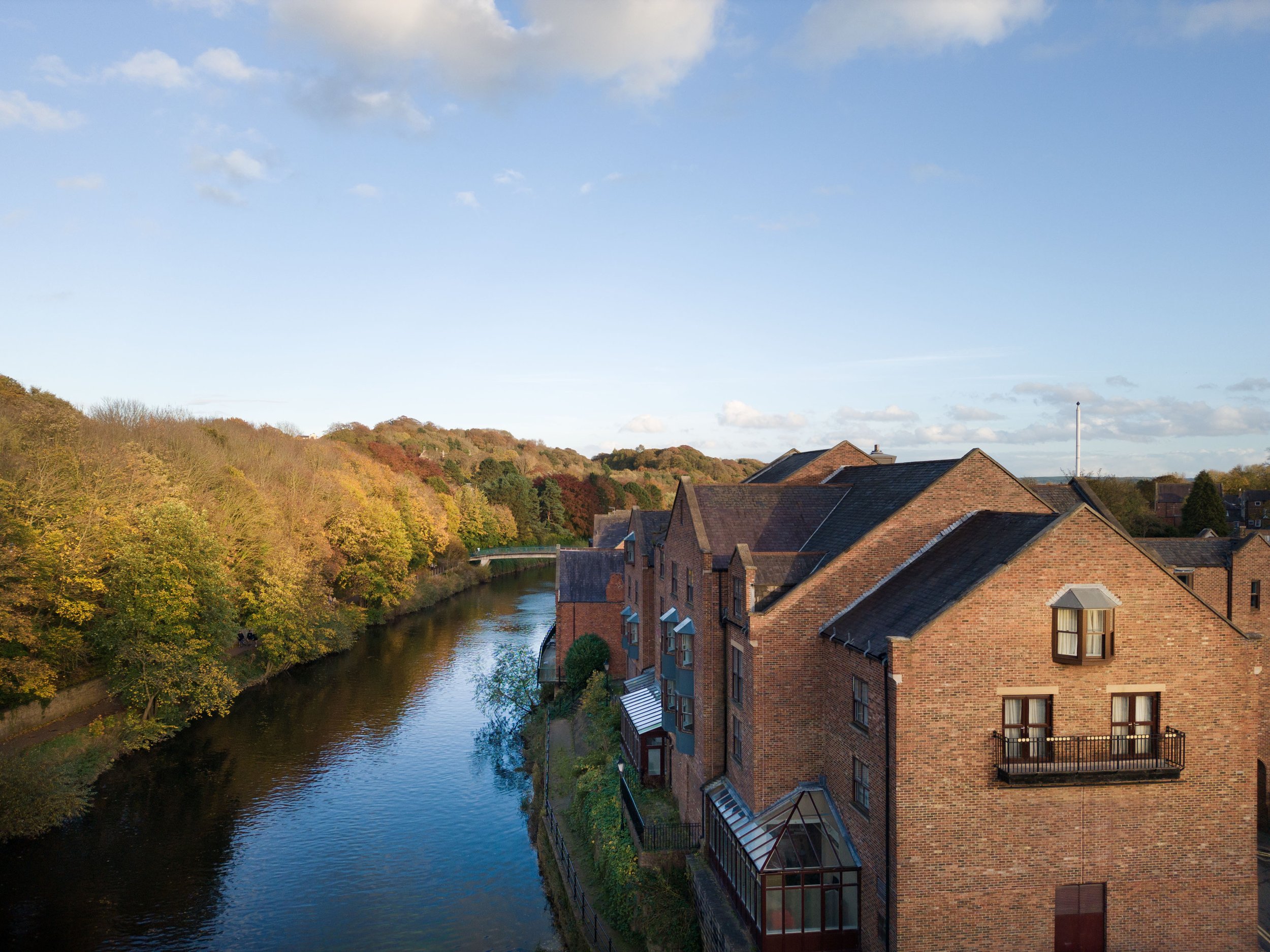A scenic view of a small river running alongside brick residential buildings with rooftops, trees with fall foliage, and a bridge in the distance under a partly cloudy sky.