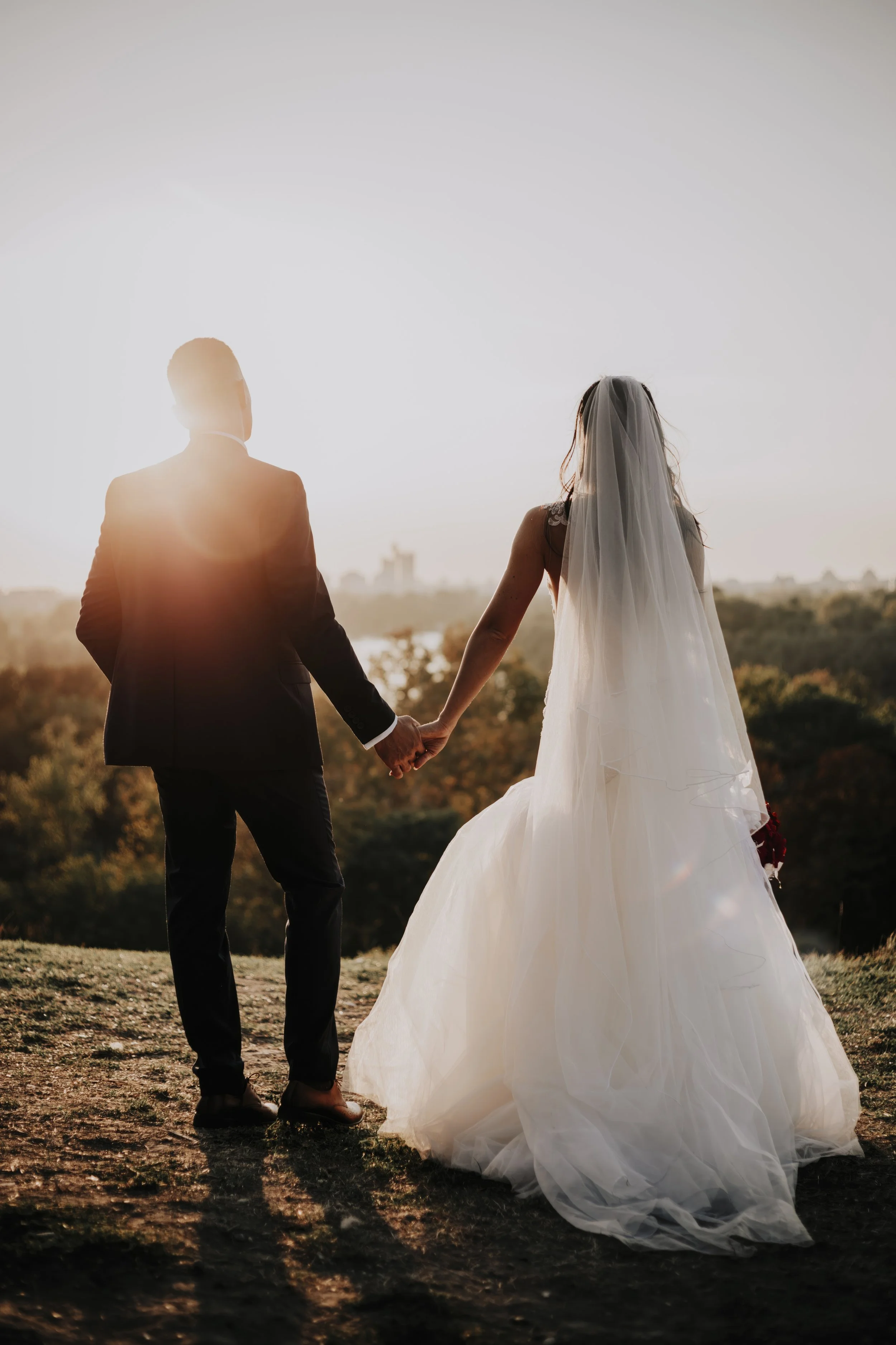 A bride and groom holding hands outdoors during sunset, with the groom in a dark suit and the bride in a white wedding gown with a veil, overlooking a landscape with trees and distant buildings.