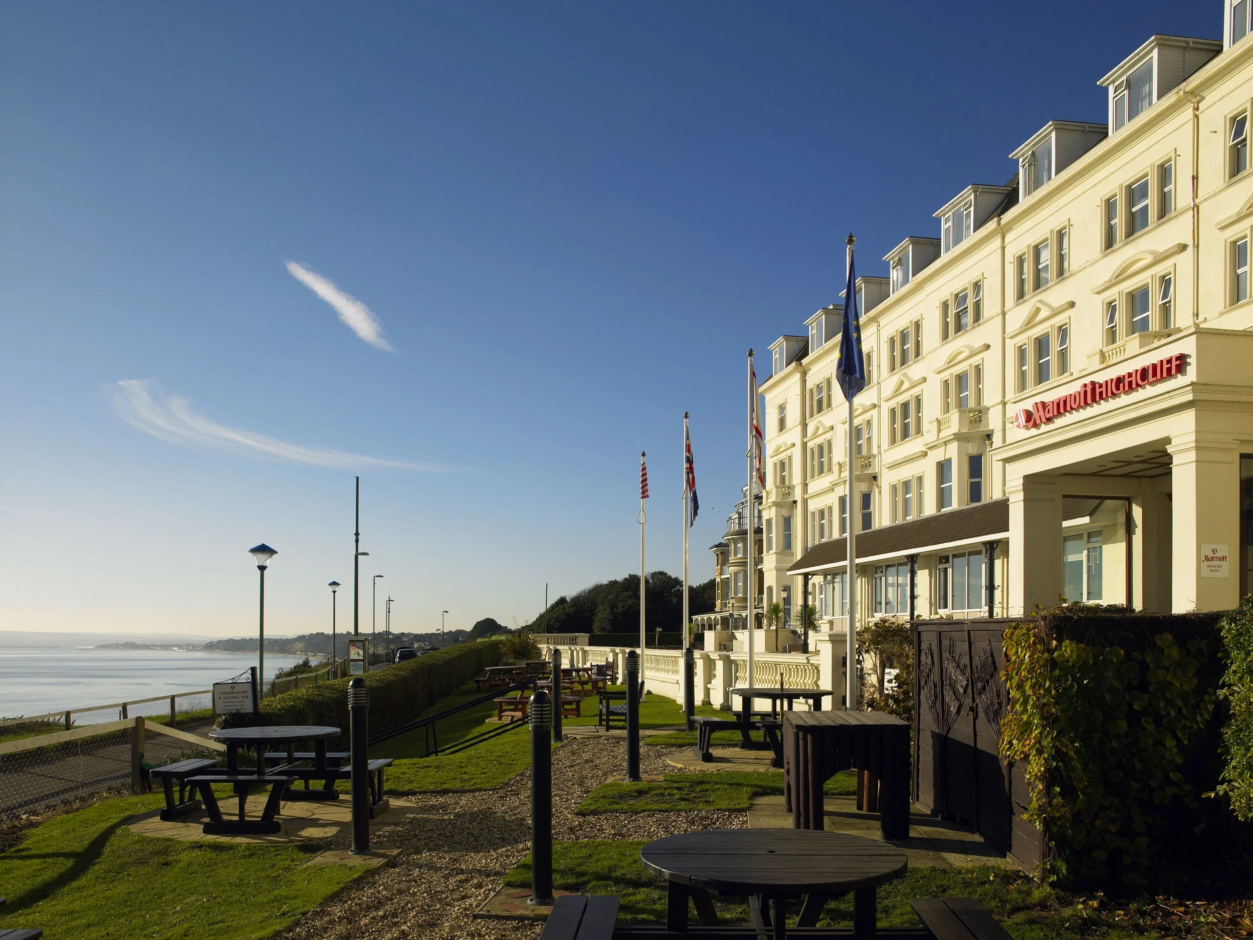 A large yellow hotel building with a Marriott Highcliff sign, flags, and multiple windows along the beachside. There are picnic tables and street lamps in the foreground, overlooking the water with a clear blue sky overhead.