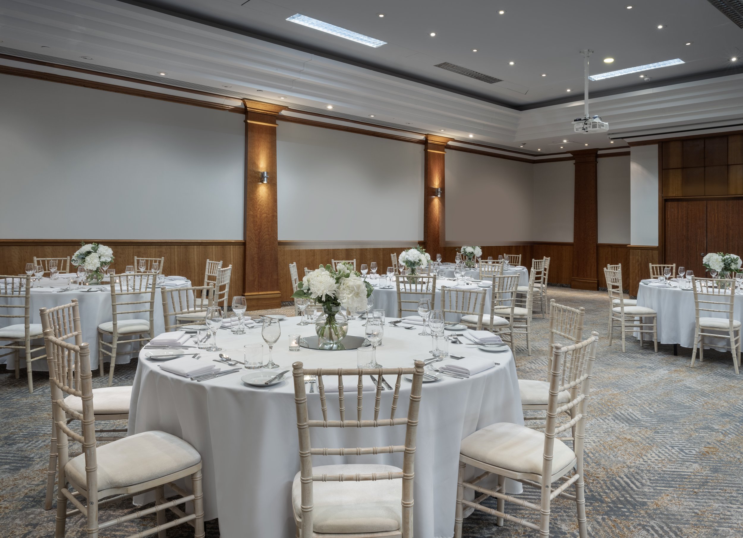 Elegant banquet hall with round tables draped in white cloths, each decorated with white floral centerpieces, surrounded by cream-colored chairs with wooden frames, and set with glassware and silverware, with a wood-paneled wall and a ceiling with recessed lighting.