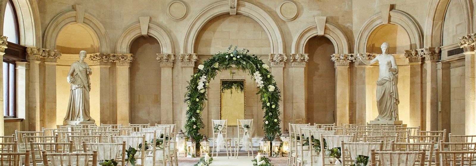 Interior of a wedding venue with rows of chairs, a floral arch, statues, and classical architecture.