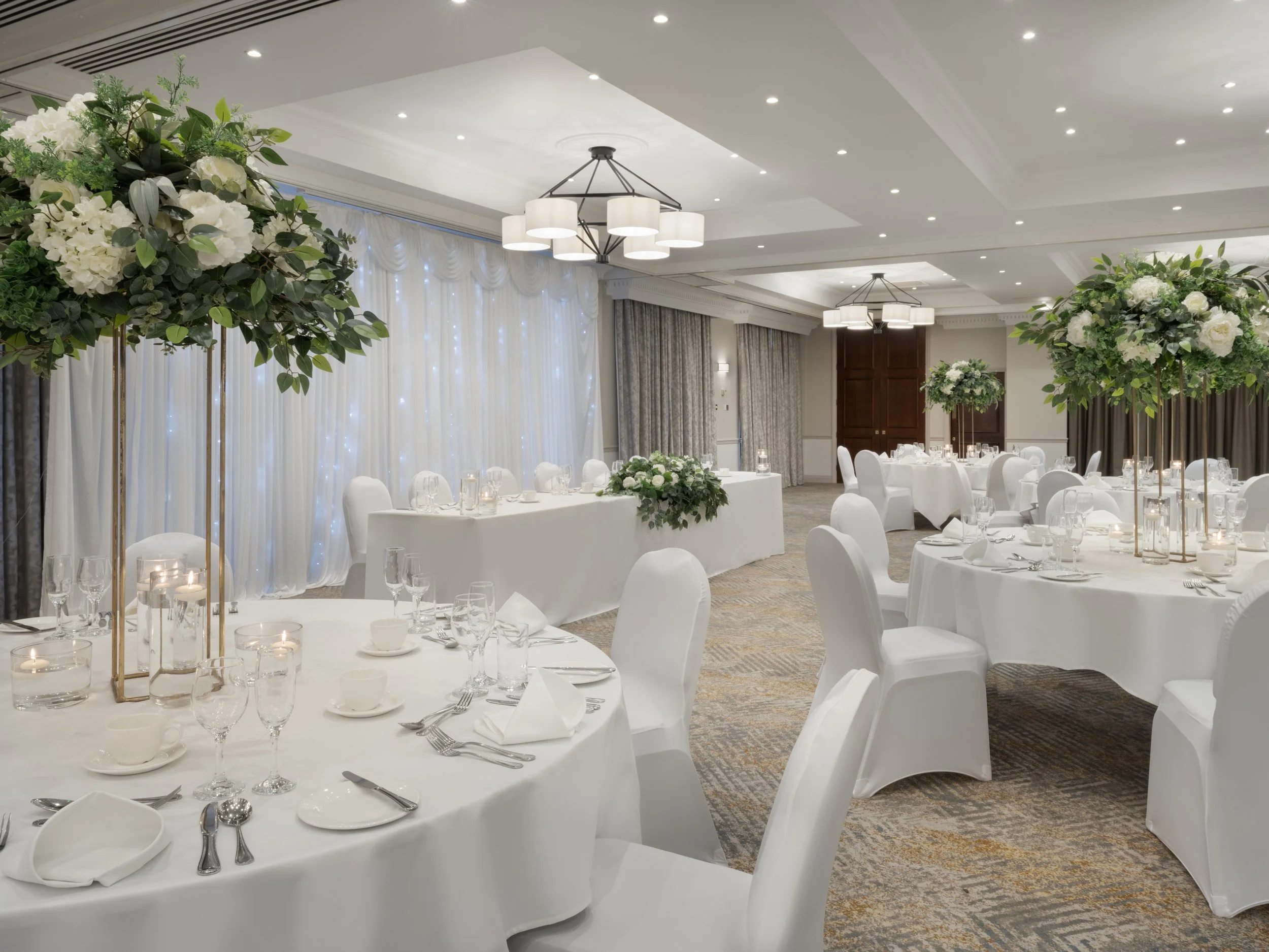 Elegant banquet hall decorated for a wedding or formal event with white tablecloths, floral centerpieces, and soft lighting.