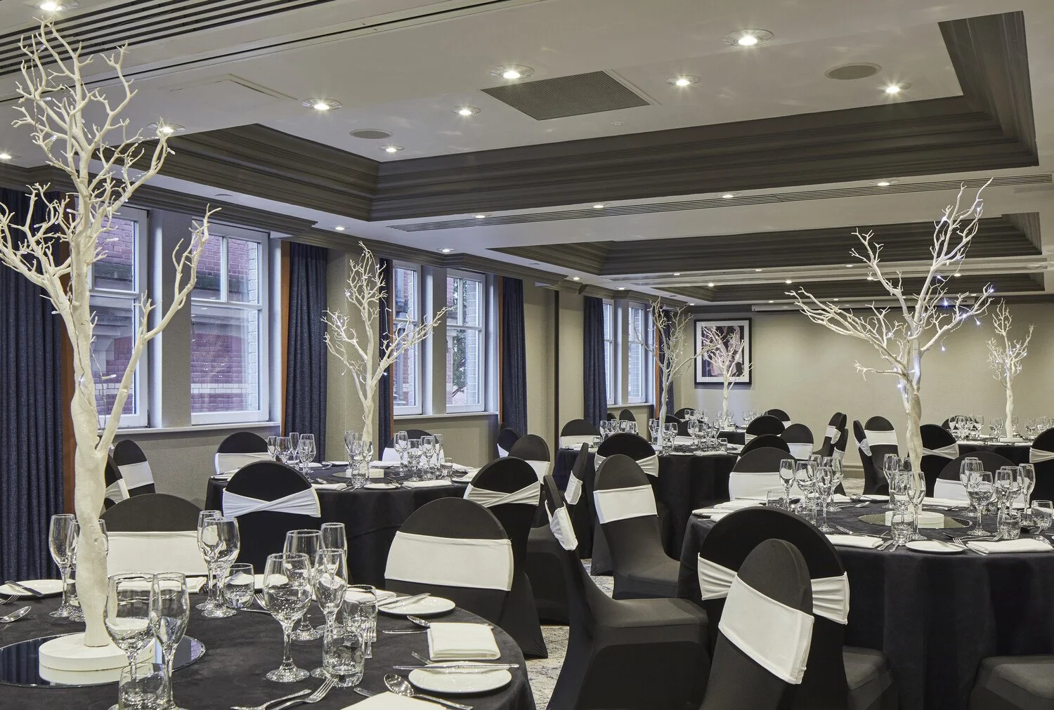 Elegant event room decorated with white leafless trees as centerpieces, black tables with black and white chairs, set with glassware and silverware, large windows with dark curtains, ceiling with recessed lighting.