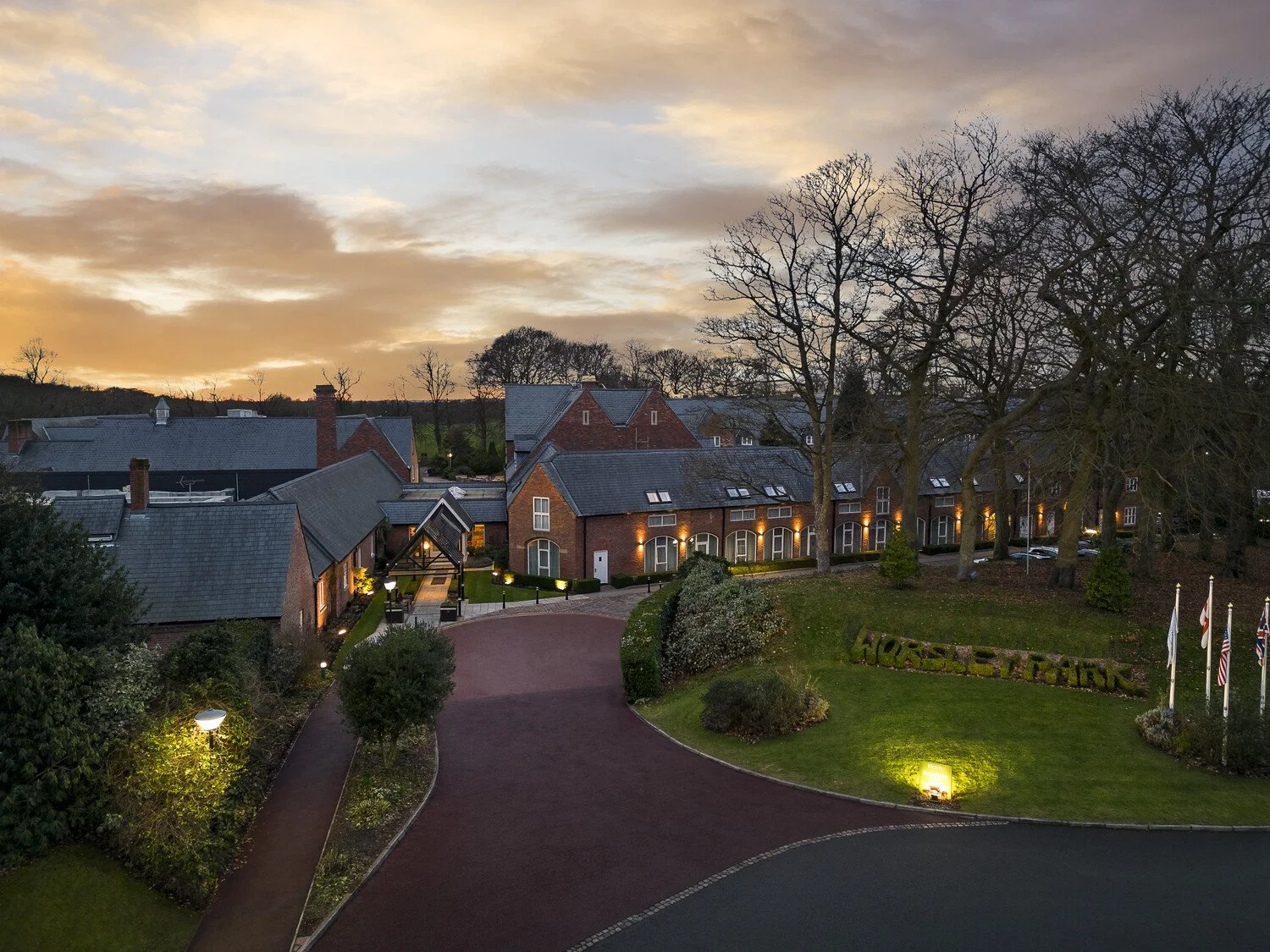 View of a hotel named Horse & Groom at sunset, with brick buildings, a curved driveway, trees, and flags in front.