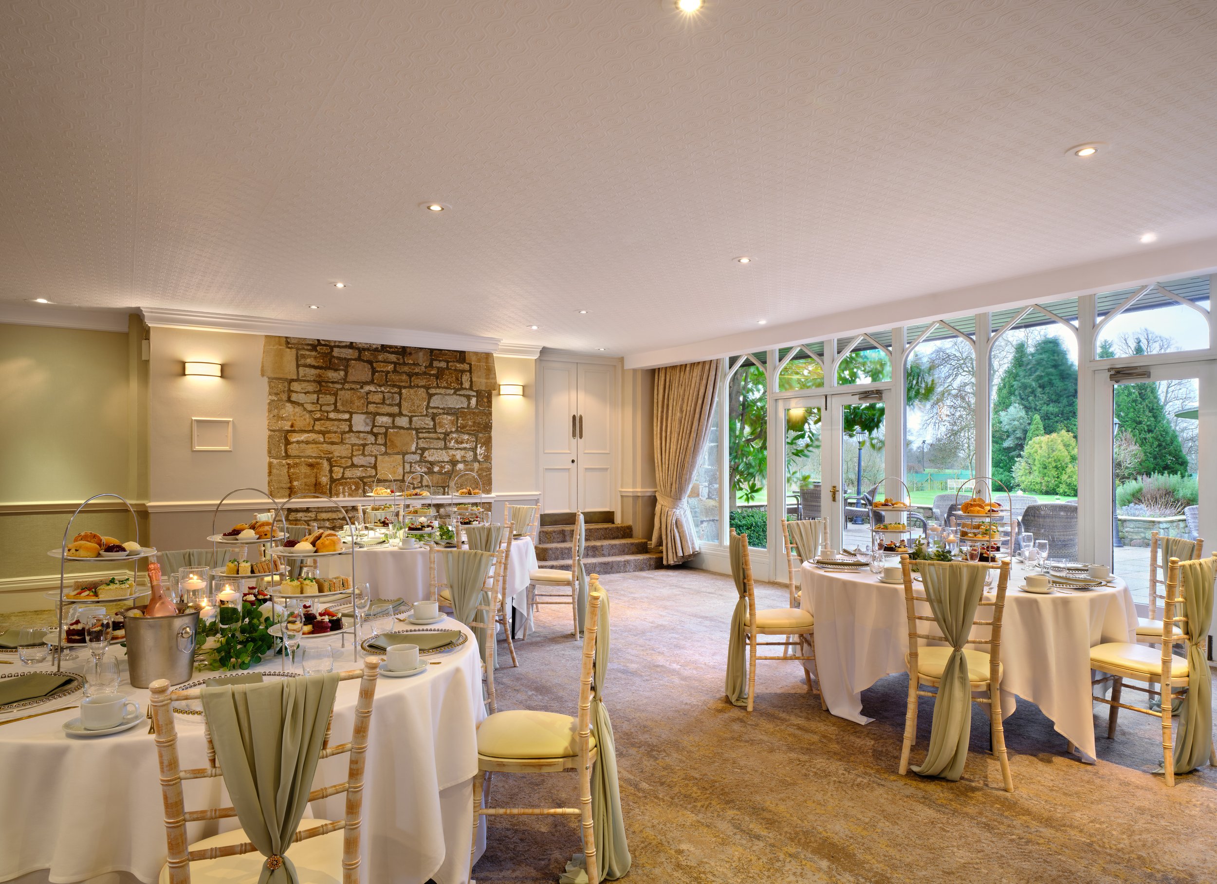 Elegant dining room set up for a formal event with round tables covered in white tablecloths, decorated with tiered trays of desserts, surrounded by chairs with cream-colored cushions, large windows revealing a scenic garden outside, stone fireplace on one wall, and soft lighting fixtures.