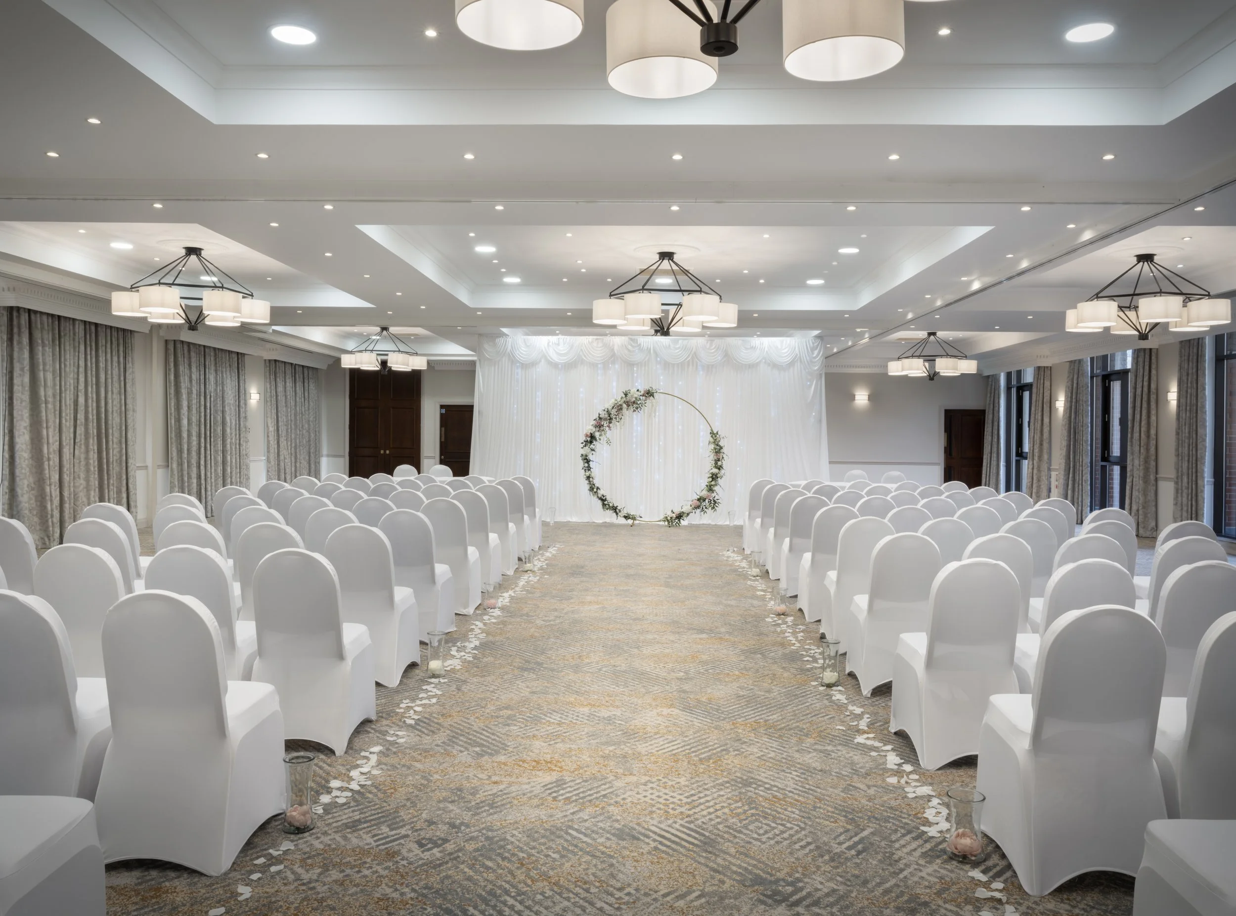 Wedding ceremony setup in a large, elegante room with rows of white covered chairs, a white draped backdrop with floral arch, and hanging chandeliers.