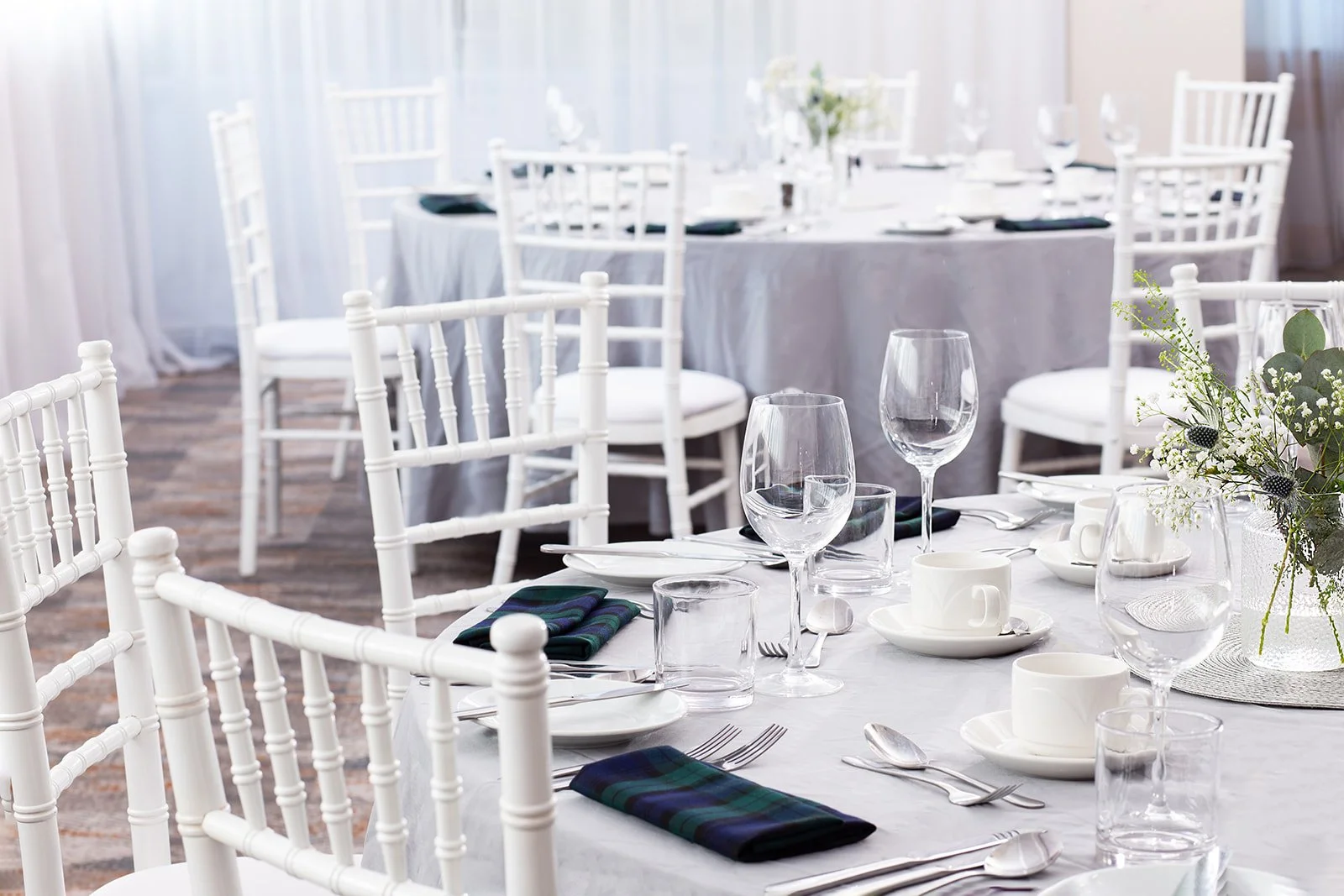 Elegant event dining setup with white chairs, a long table, white tablecloth, wine glasses, white cups and saucers, silverware, folded napkins, and a floral centerpiece in a well-lit room with sheer curtains.