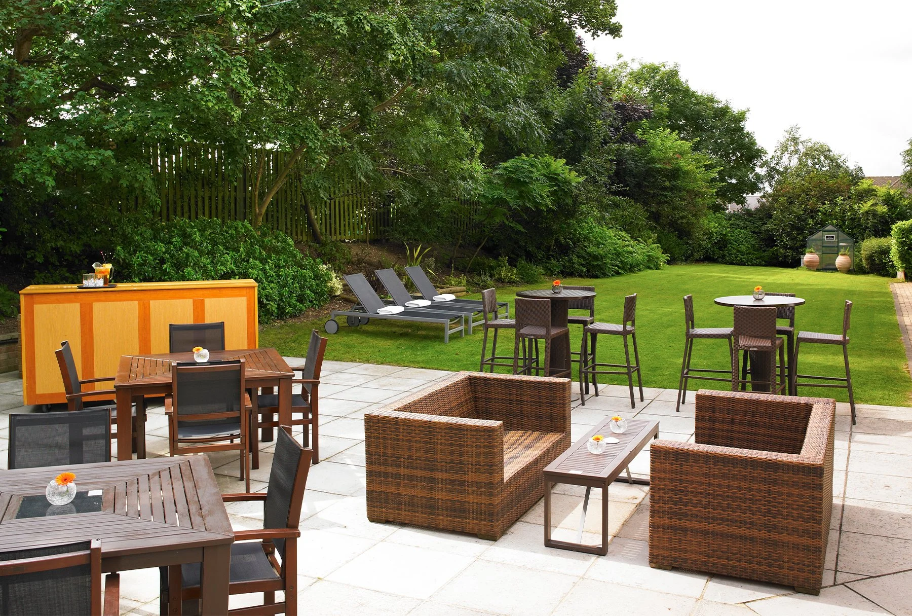 Outdoor patio with tables and chairs, some with small flower vases, overlooking a lush green garden with trees and a small shed in the background.