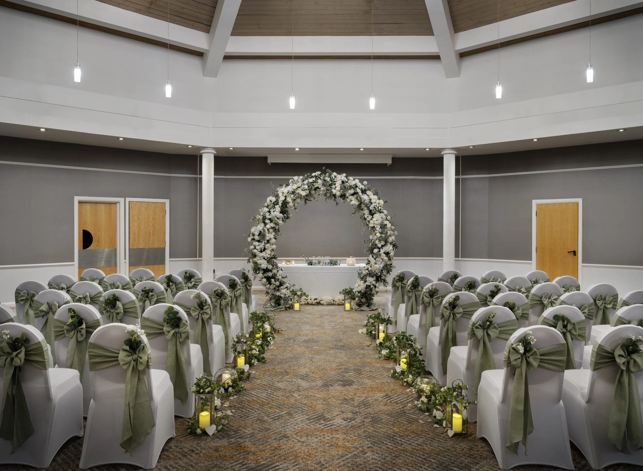 Wedding ceremony setup with rows of white chairs decorated with green ribbons and flowers, a floral arch, and aisle candles in a spacious hall with a high ceiling.