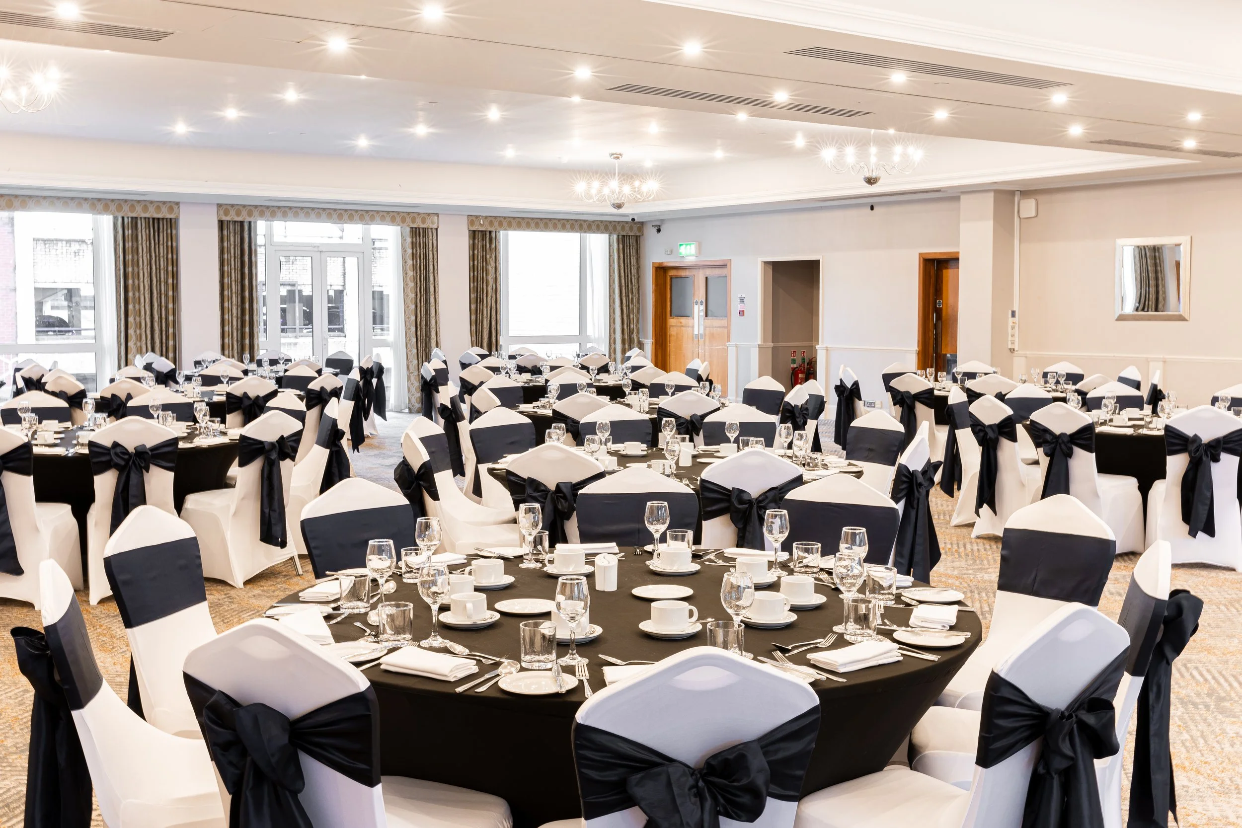 A banquet hall with round tables covered with black tablecloths, set with white plates, cups, and glassware, surrounded by chairs with white covers and black bows. The room has large windows, chandeliers on the ceiling, and a beige carpeted floor.