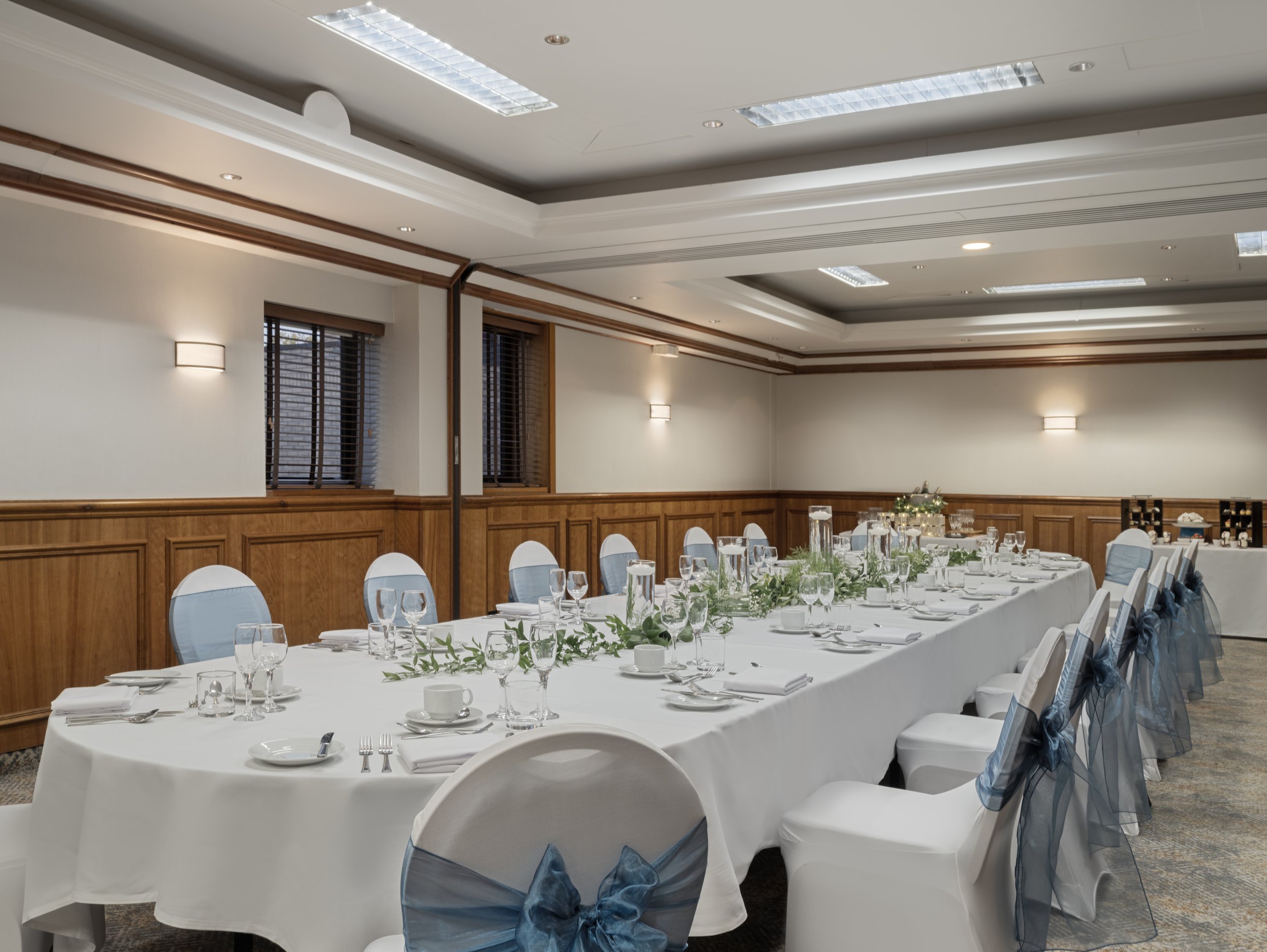 A banquet hall with tables set for a formal event, decorated with white tablecloths, glassware, and greenery, with chairs covered in white fabric and adorned with blue bows, and a ceiling with recessed lighting and wood paneling.
