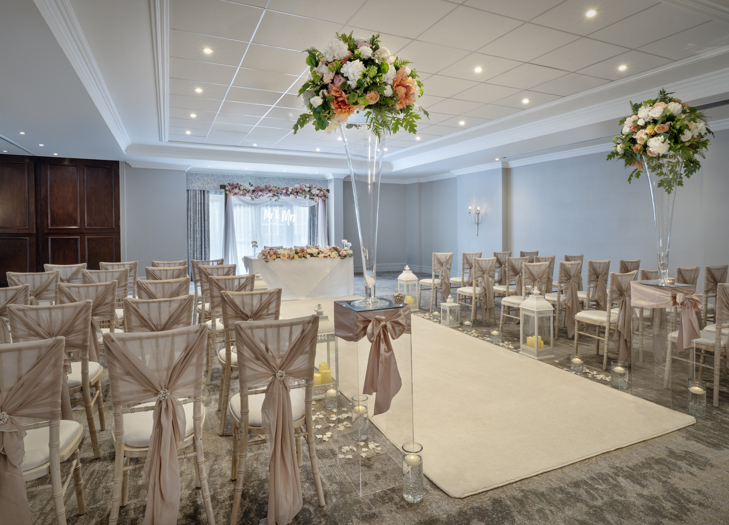 A wedding ceremony setup with rows of chairs decorated with pink bows, floral arrangements on tall stands, and a decorated table at the front with a "Mr & Mrs" sign in the background.