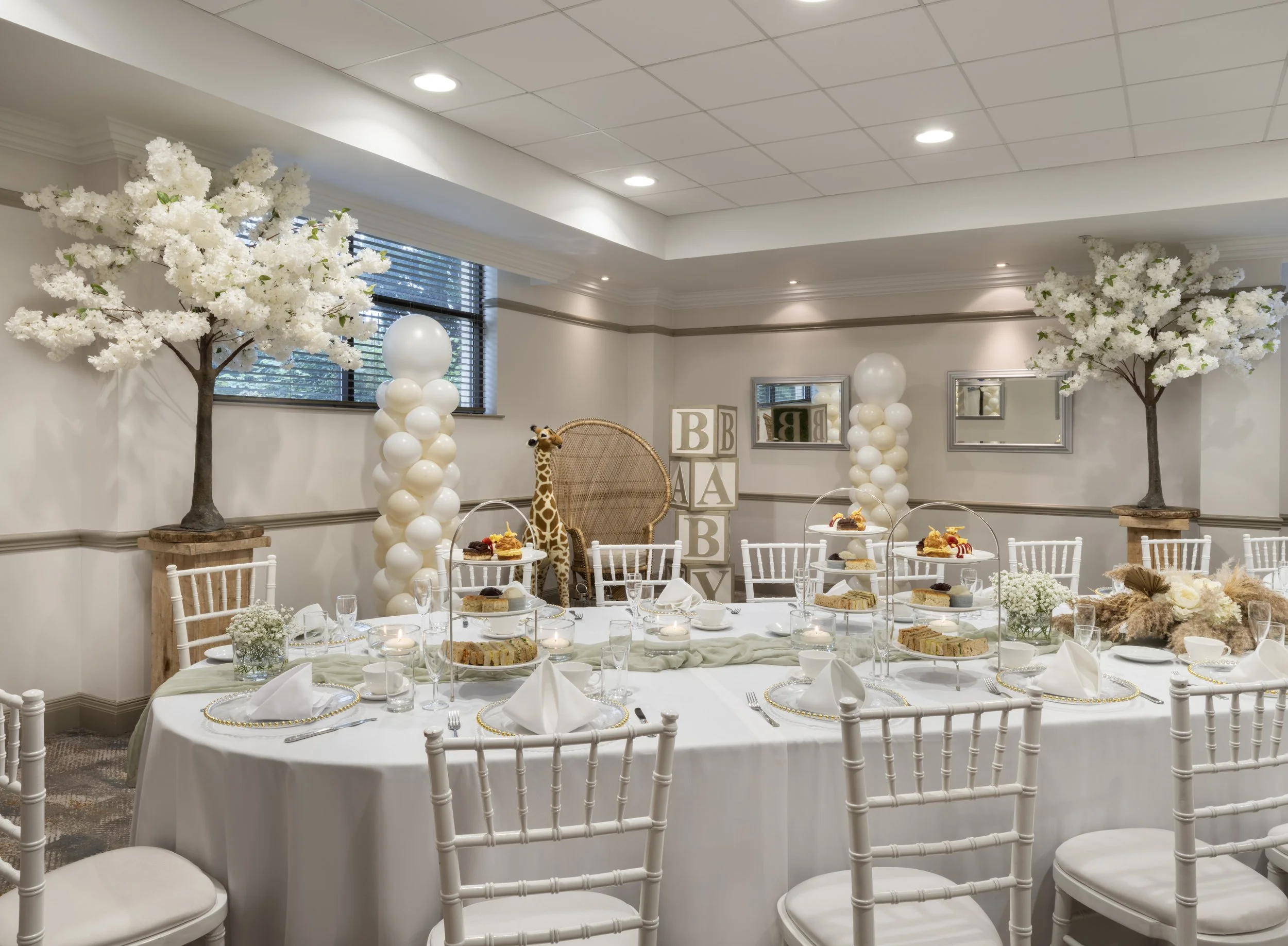 Decorated banquet room with white floral trees, balloon columns, a giraffe plush toy, framed alphabet blocks, and a dining table set with white dinnerware and desserts.