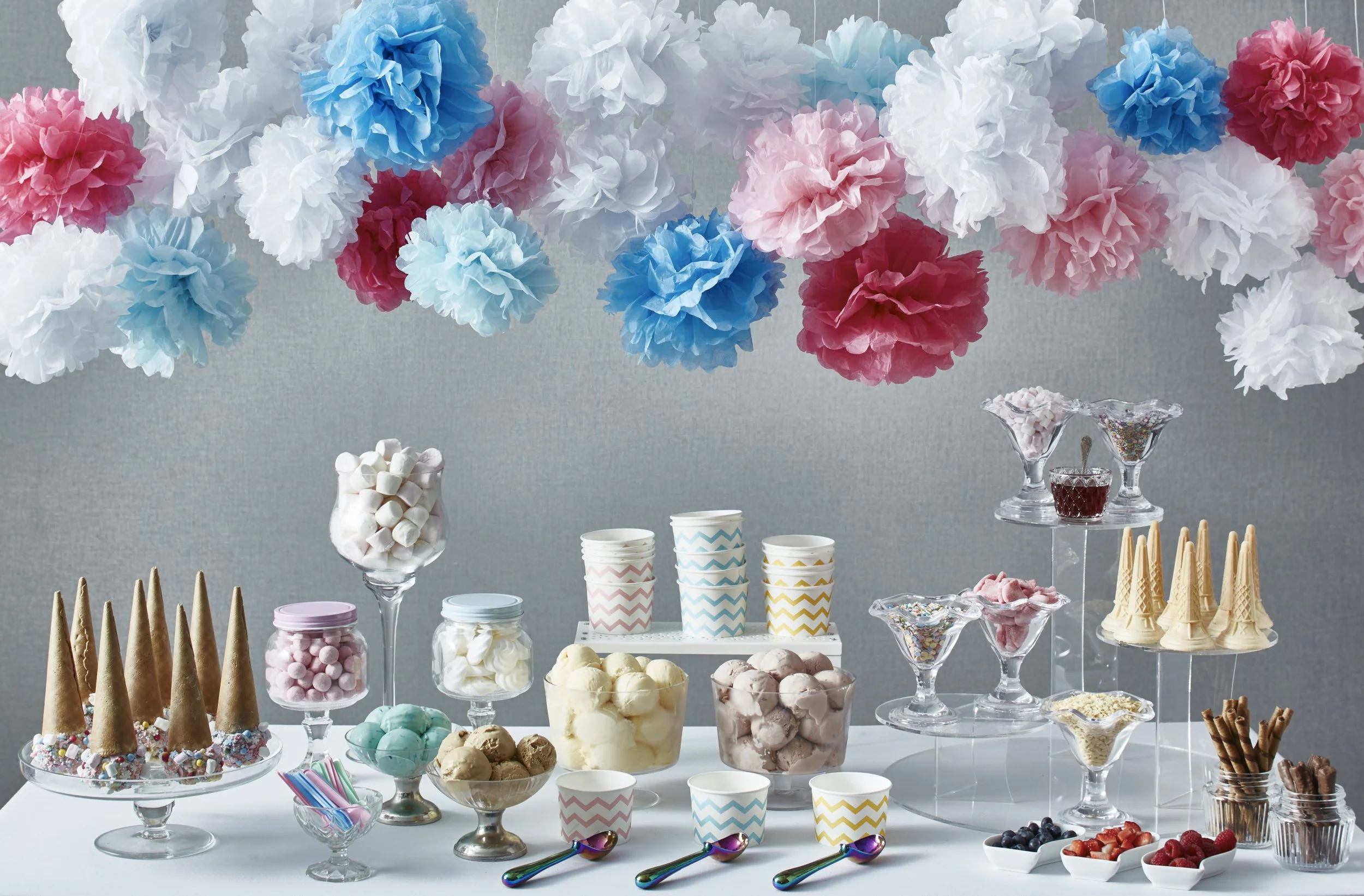 A colorful dessert table with various treats including ice cream, candies, fruit, and cones, decorated with tissue paper pom-poms in pink, blue, and white hanging above.