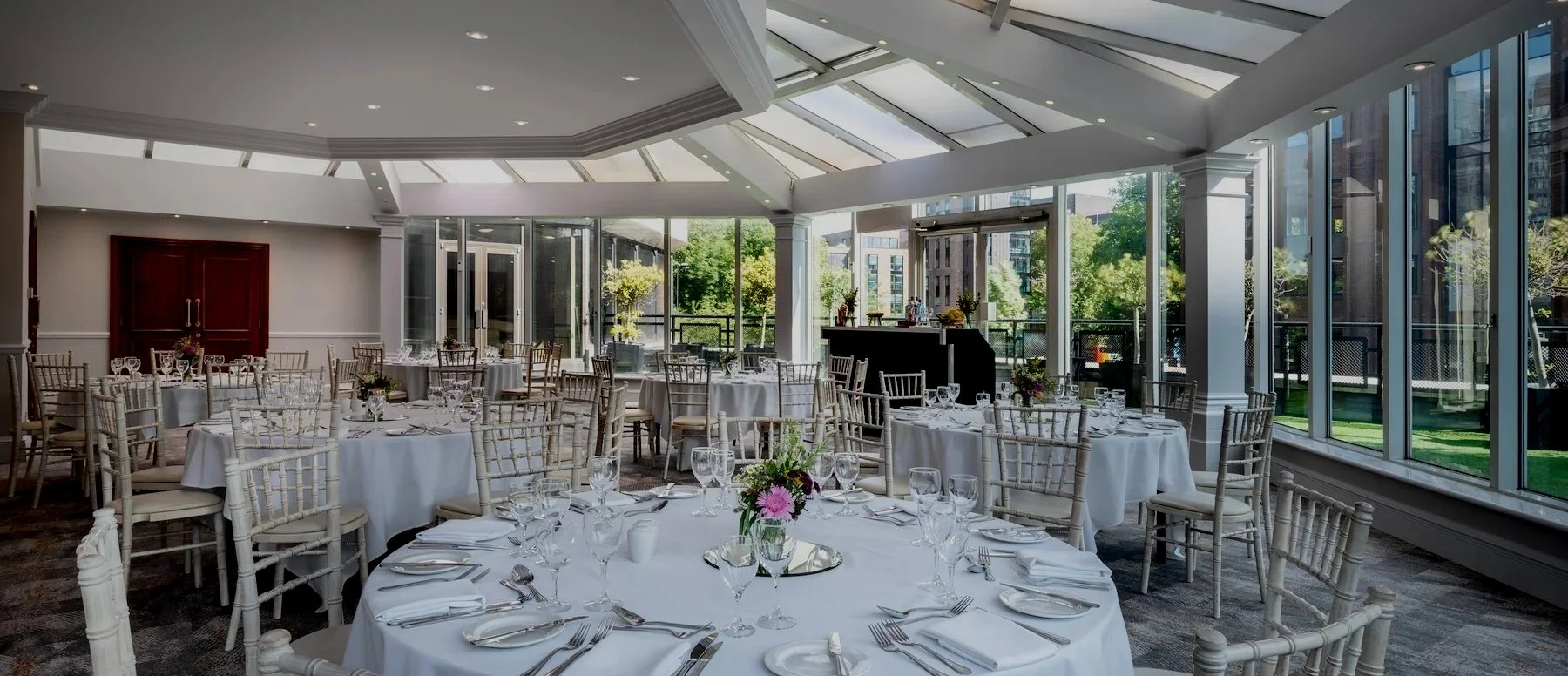 Elegant event space with round tables covered in white tablecloths, set with glassware, silverware, and floral centerpieces. Large windows and a glass ceiling let in natural light, with a view of trees outside.